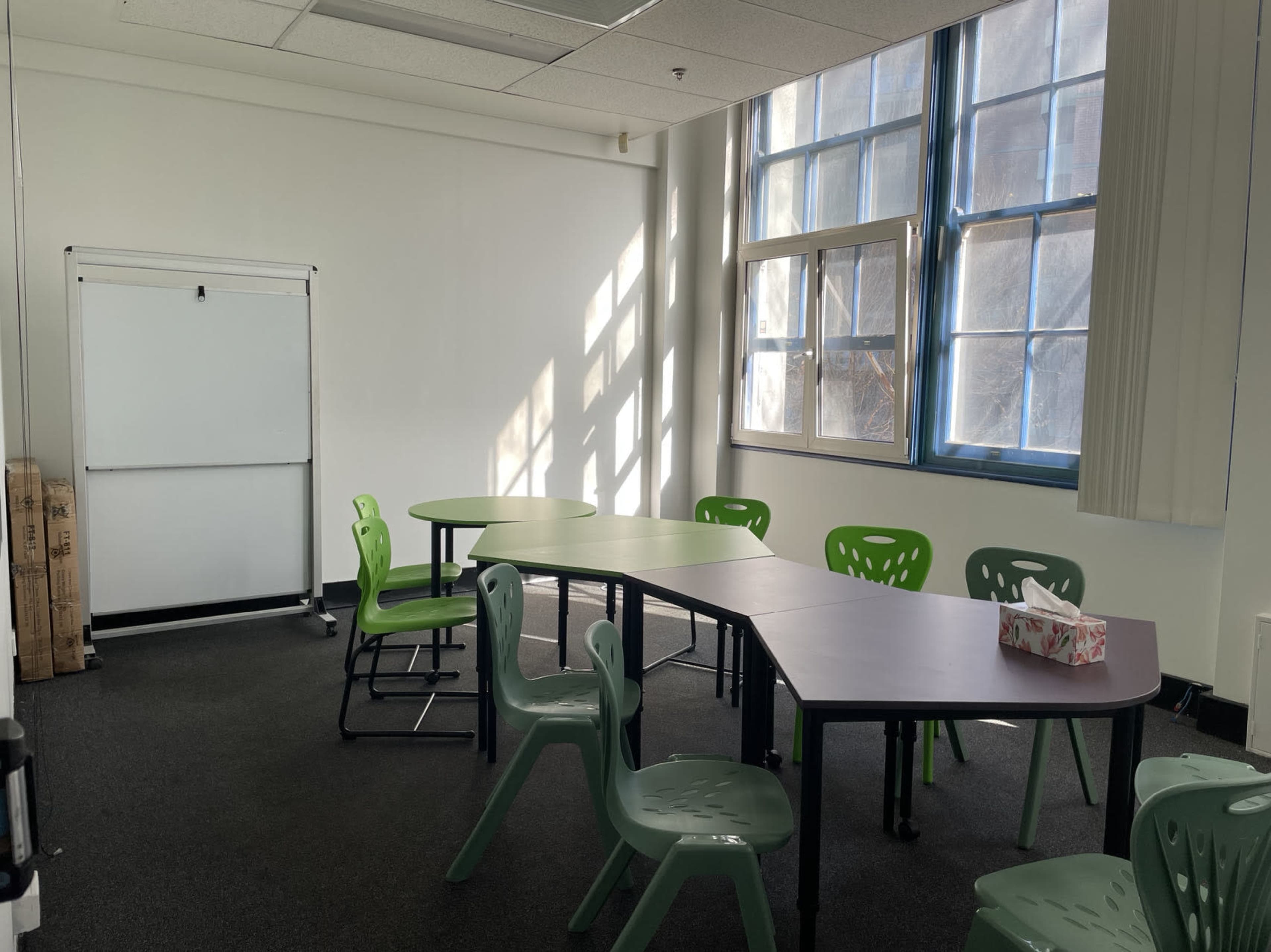A small classroom with green chairs and tables arranged in a U-shape, featuring a whiteboard and large windows allowing sunlight to enter.