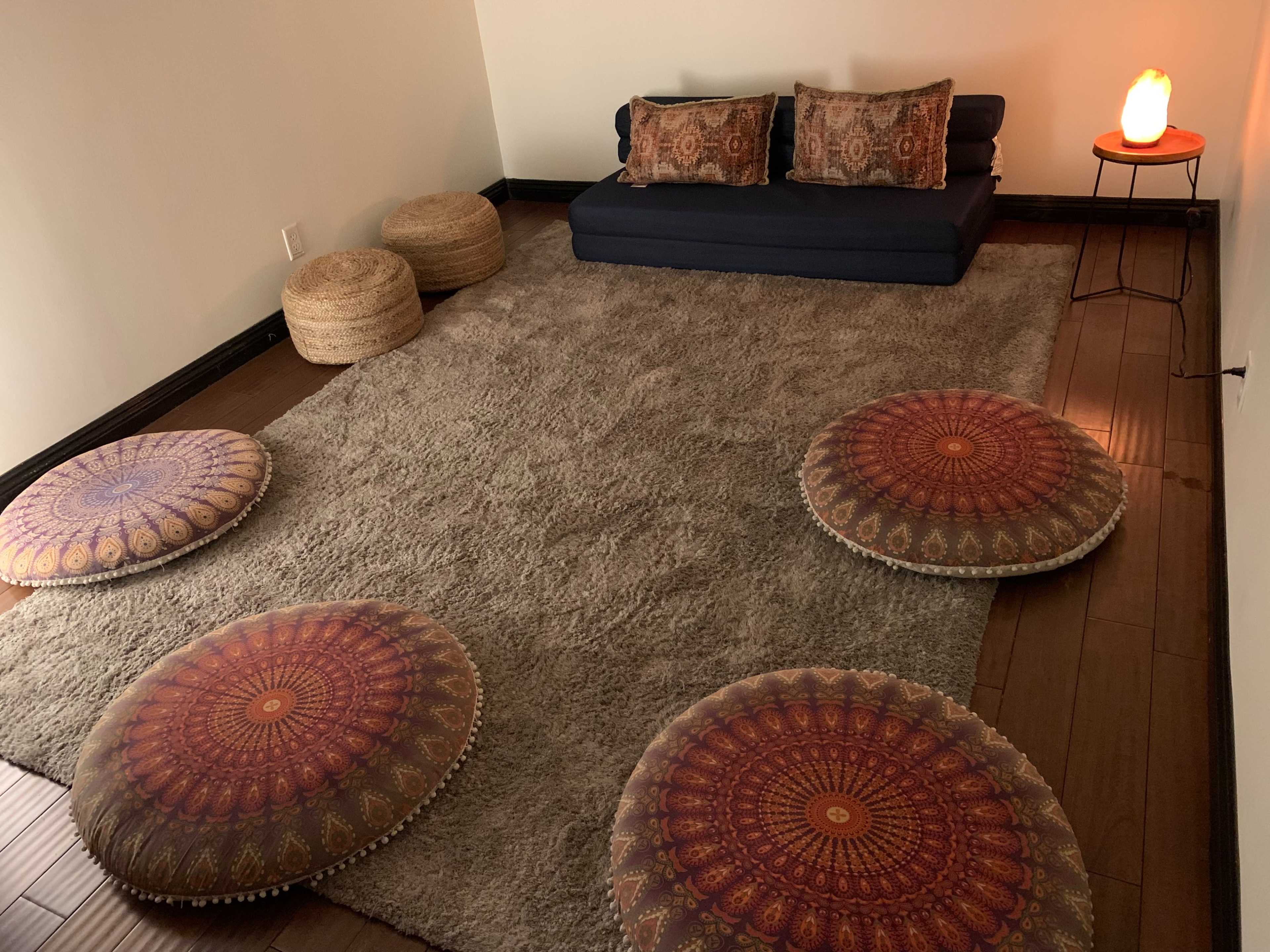 The image shows a cozy, minimalist meditation space featuring a dark sofa, round patterned cushions, a lamp, and a soft, textured rug.