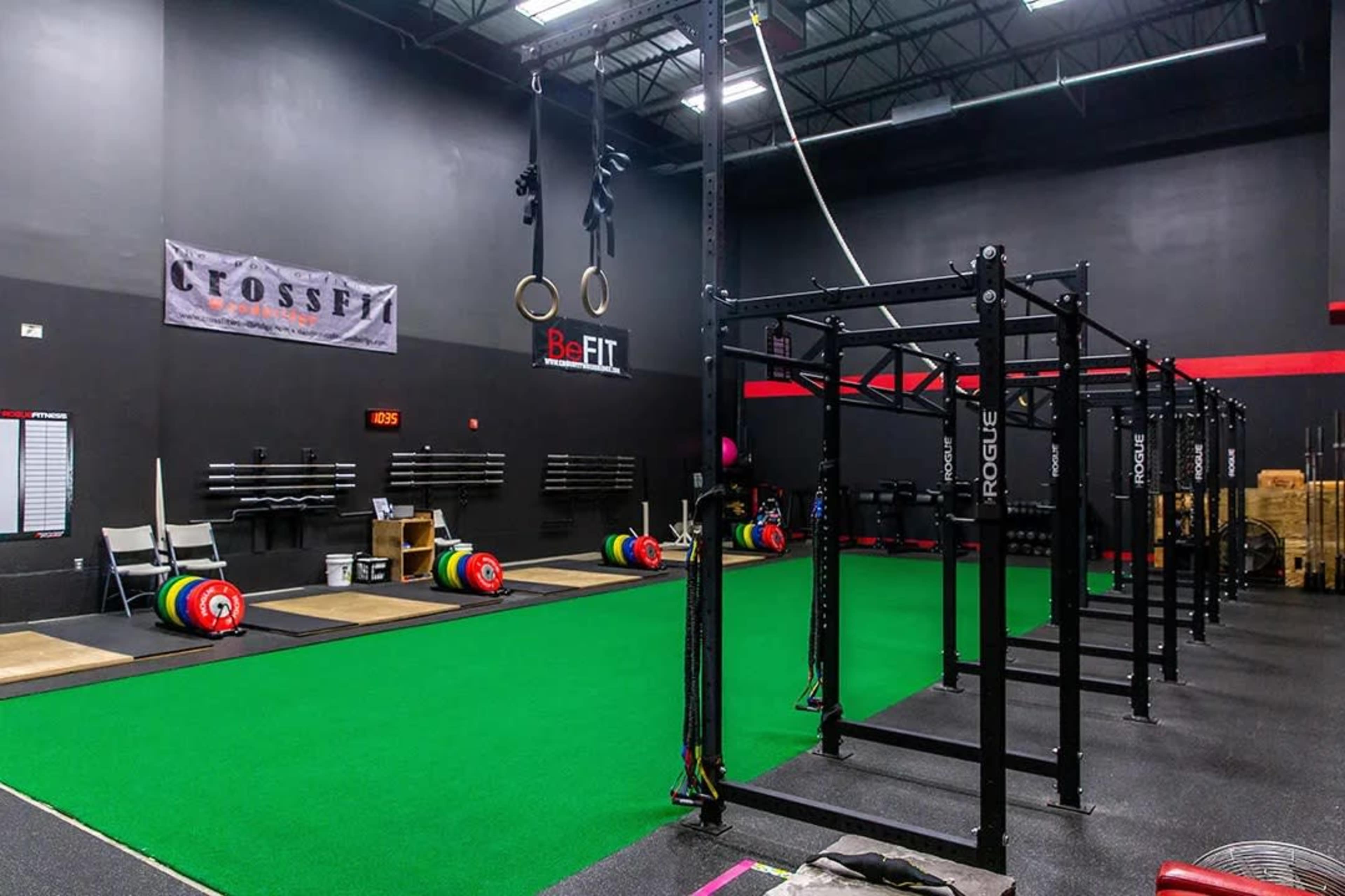 A CrossFit gym featuring a green turf area, weightlifting platforms, and a structure for rings and pull-ups, with various barbells and weights stored nearby.
