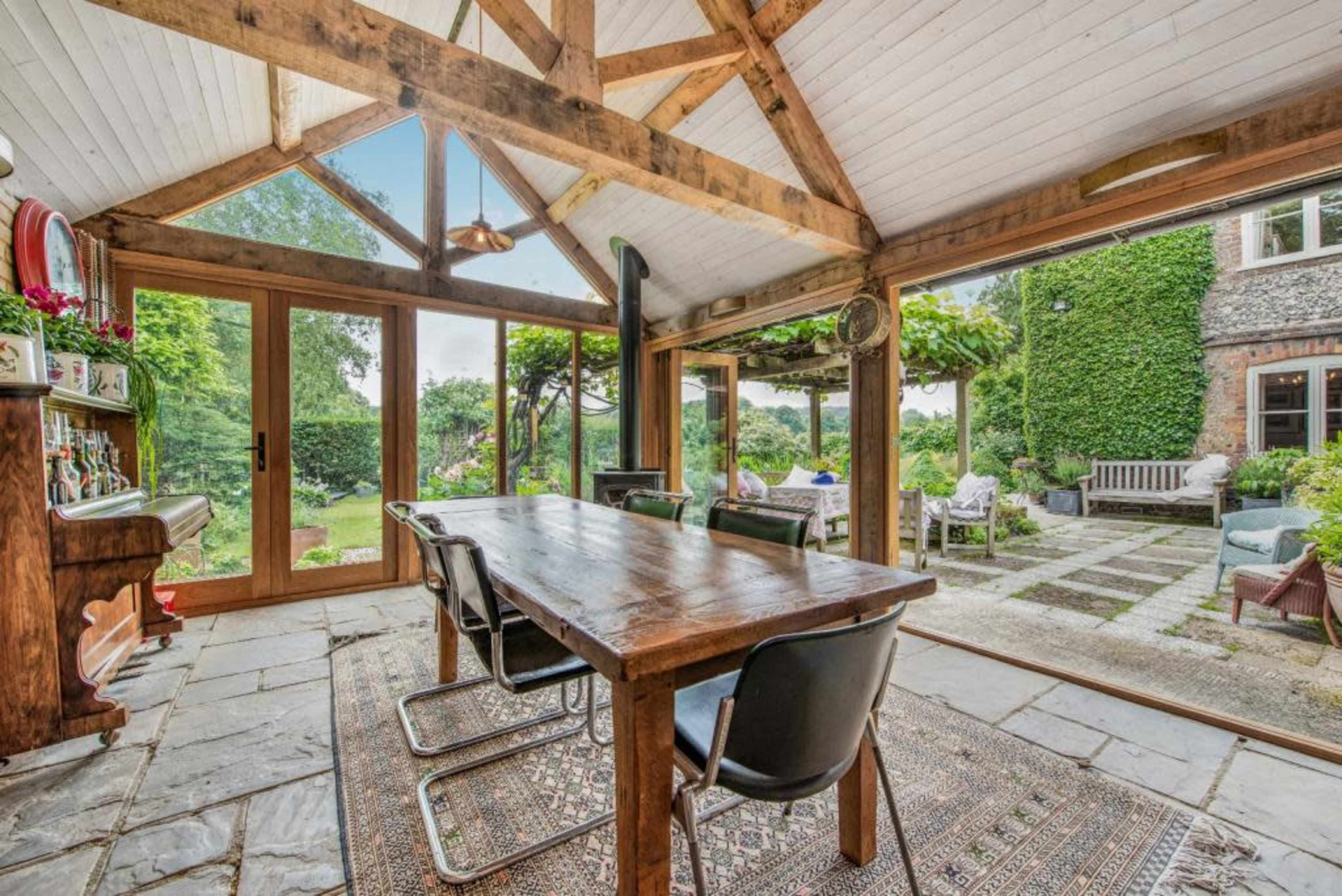 The image shows a wooden dining table with black chairs in a glass-enclosed room that opens to a garden, featuring stone flooring and a rustic ceiling.