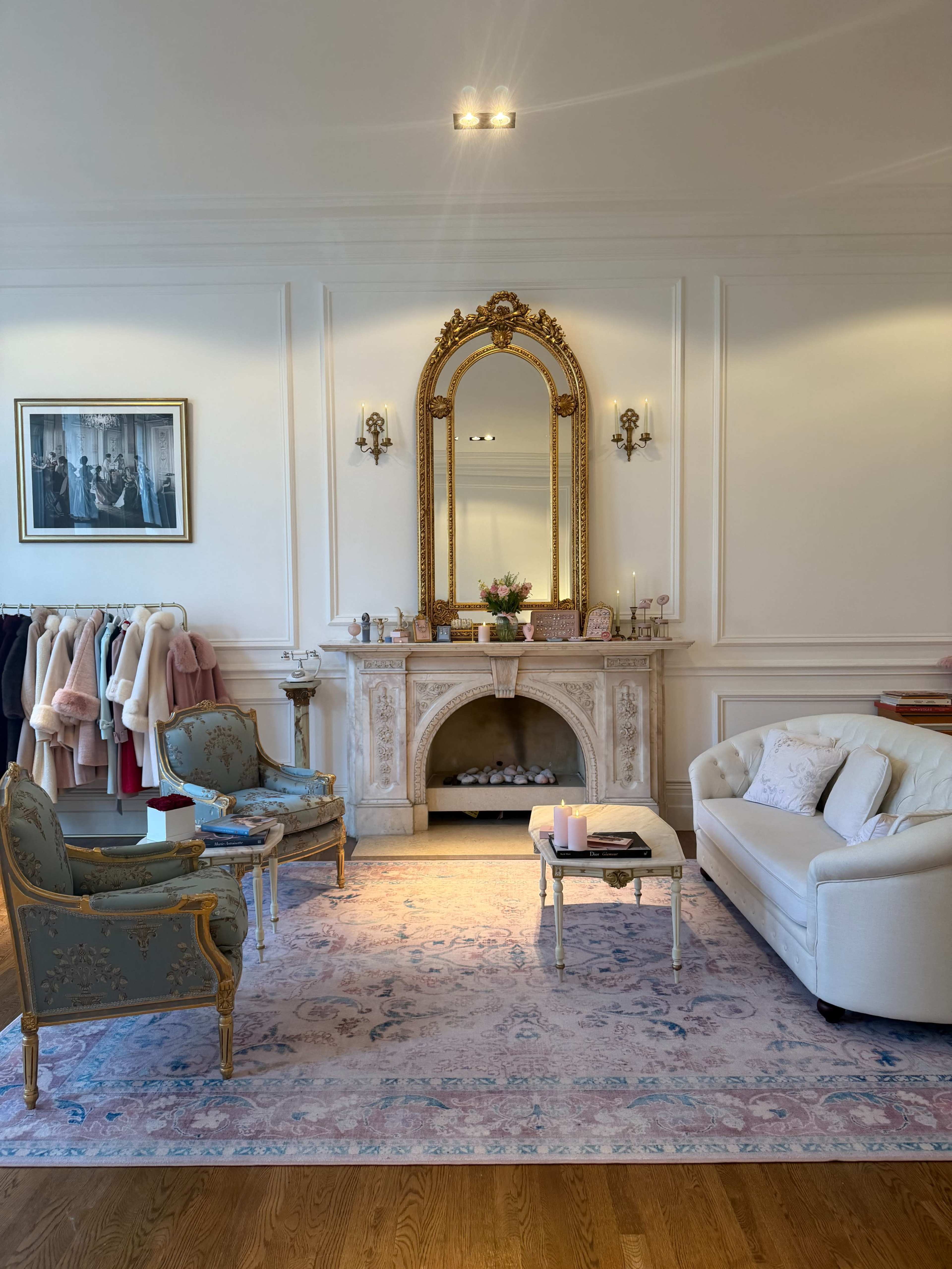 The image shows an elegantly styled living room featuring a marble fireplace, a large mirror, a white sofa, and ornate chairs, adorned with various clothing items on display.