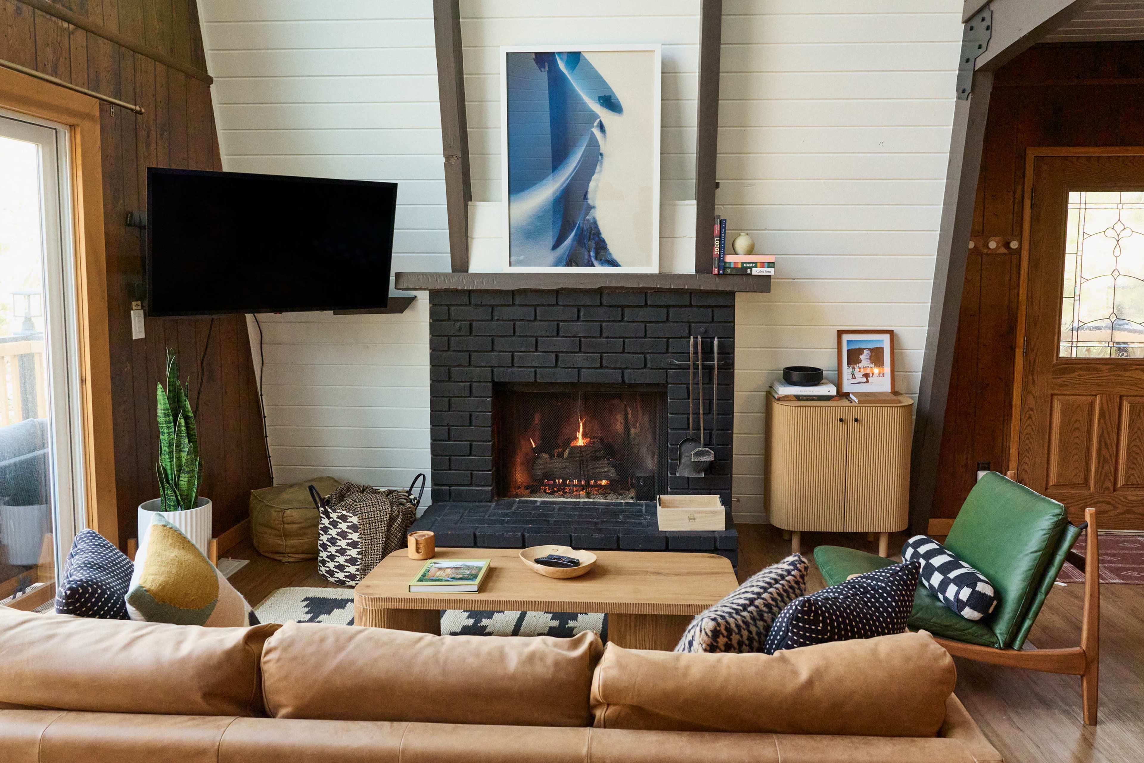 The image shows a cozy living room with a modern sofa, a coffee table, a stone fireplace, and a large wall-mounted TV, featuring wooden walls and a bright artwork above the fireplace.