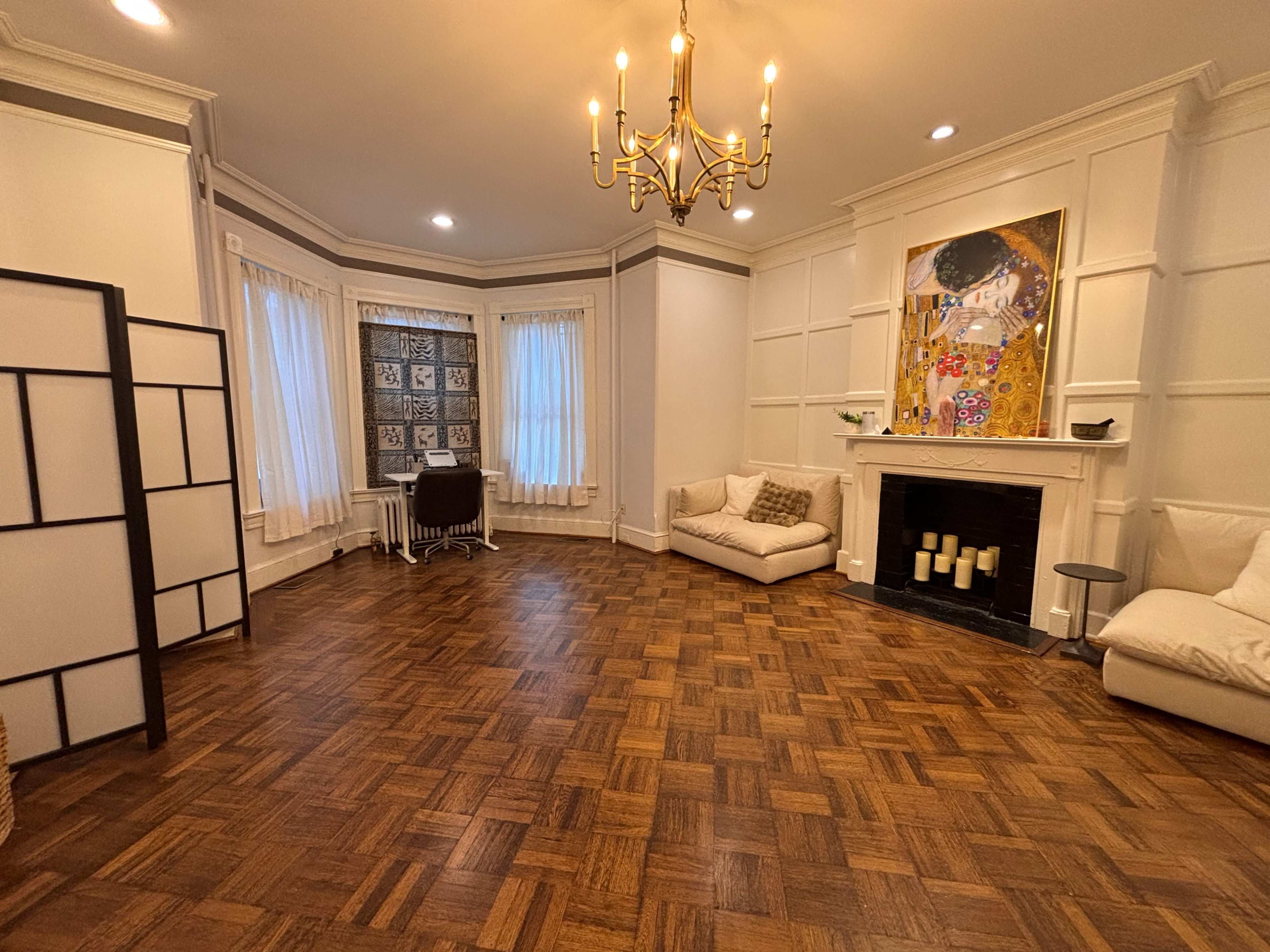 The image shows a spacious living room with a patterned wood floor, large windows dressed in sheer curtains, a white couch, and a fireplace featuring artwork above it.