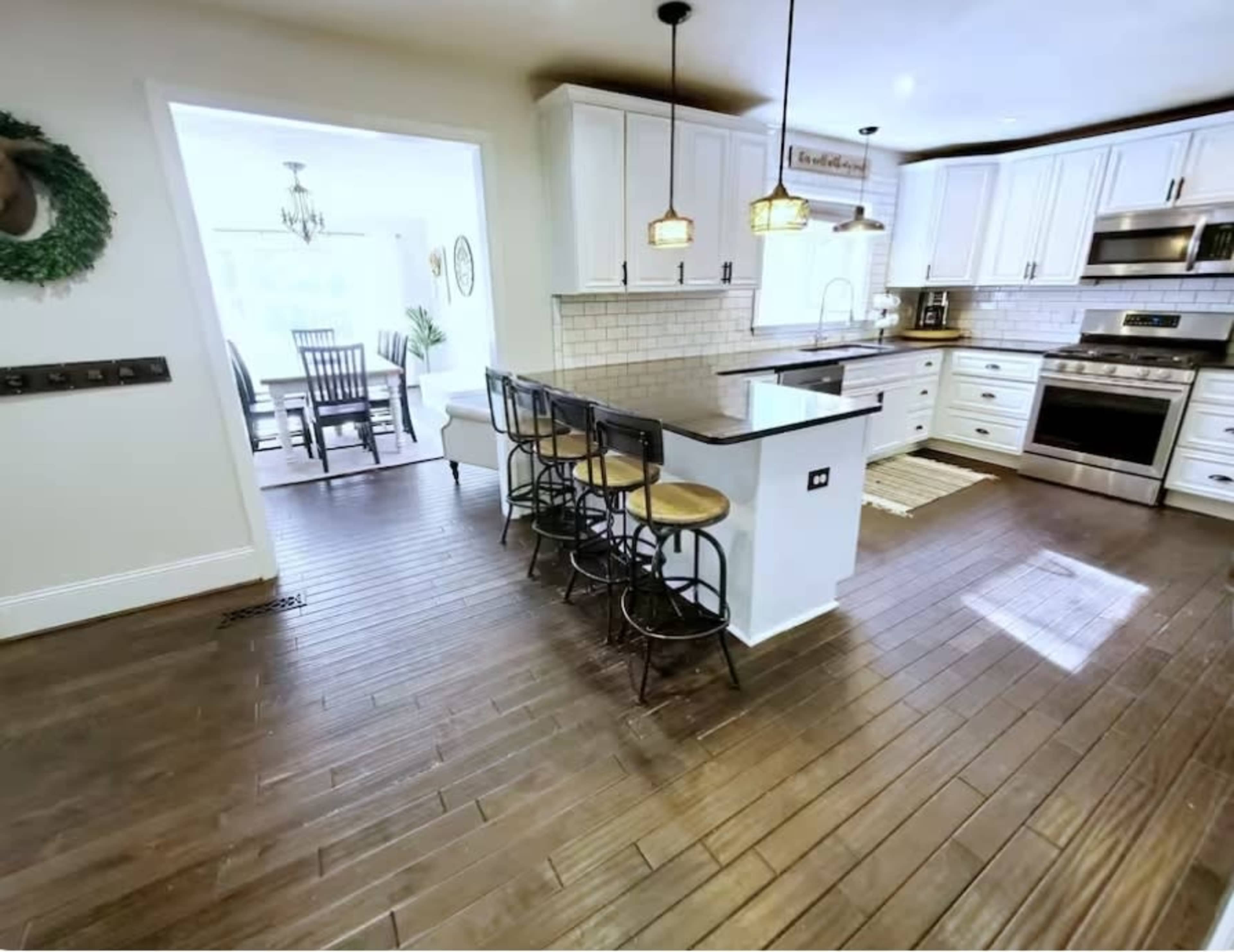 The image shows a modern kitchen with wooden floors, white cabinetry, an island with bar stools, and a dining area visible in the background.