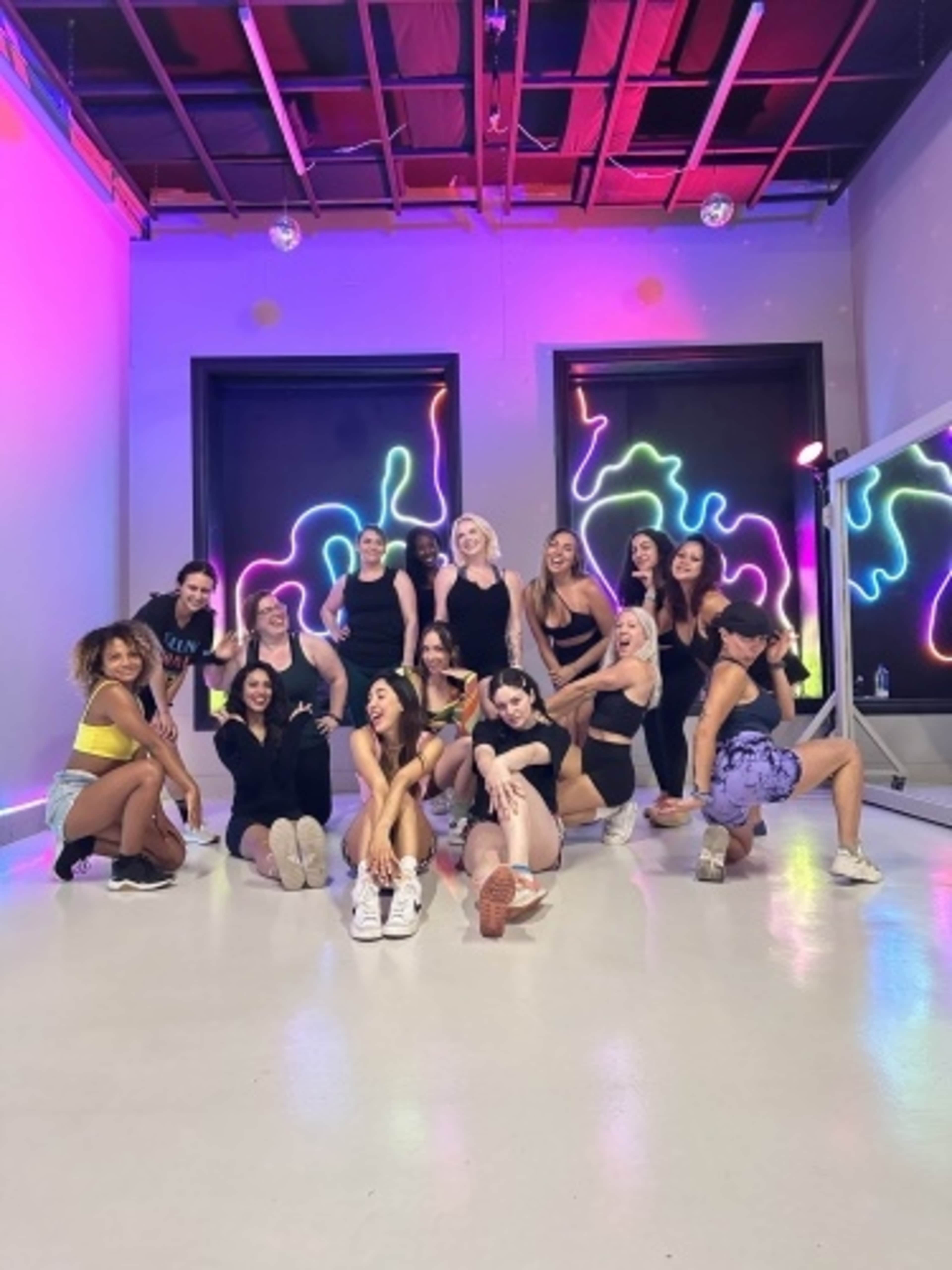 A group of young women poses in a brightly lit dance studio with colorful neon artwork on the walls.