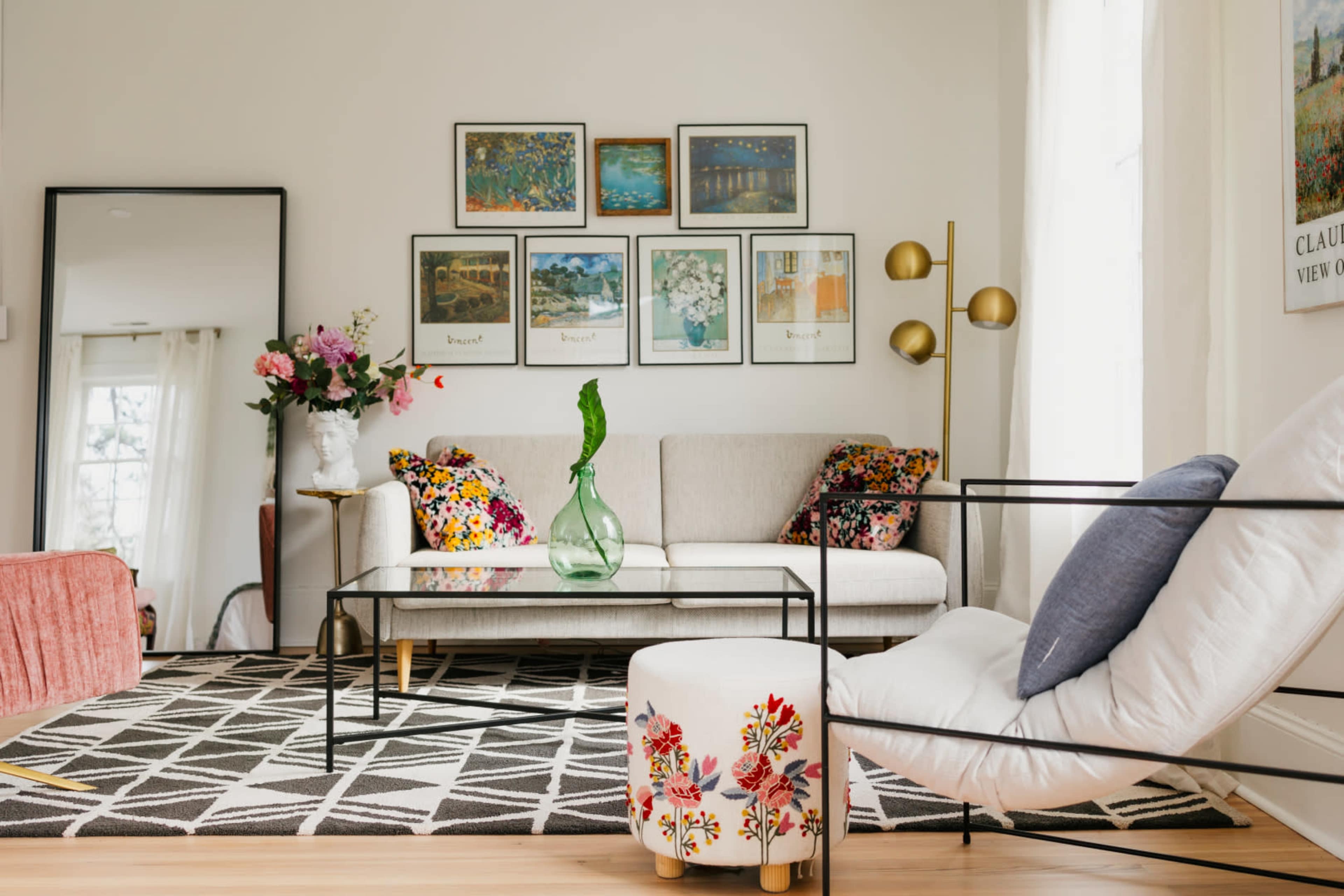 The image shows a modern living room with a sofa, coffee table, decorative pillows, and framed artwork on the wall, complemented by a floor mirror and a vase of flowers.