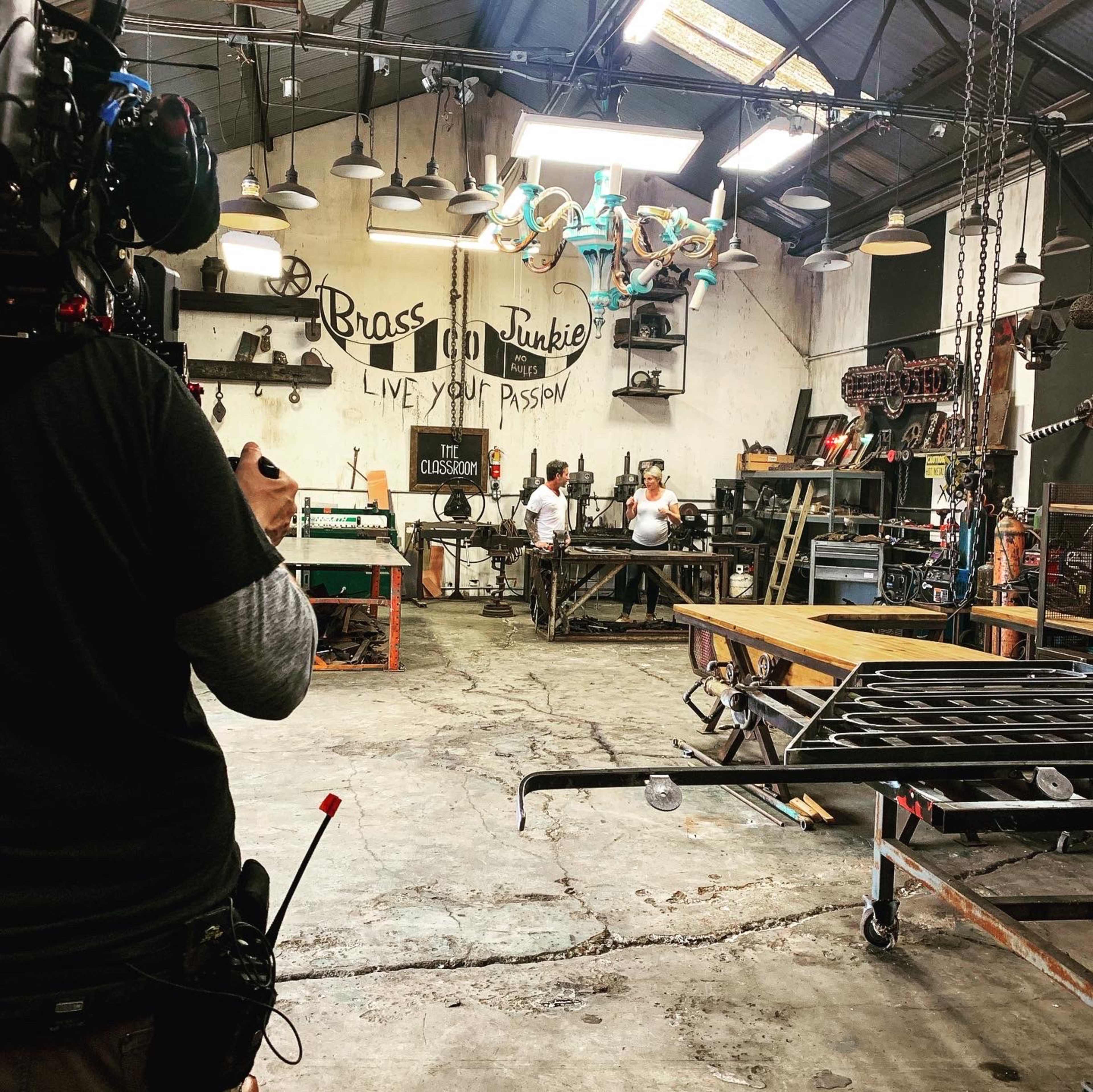 A person operates a camera while two individuals work with metal tools in a cluttered workshop filled with various equipment and decorative items.