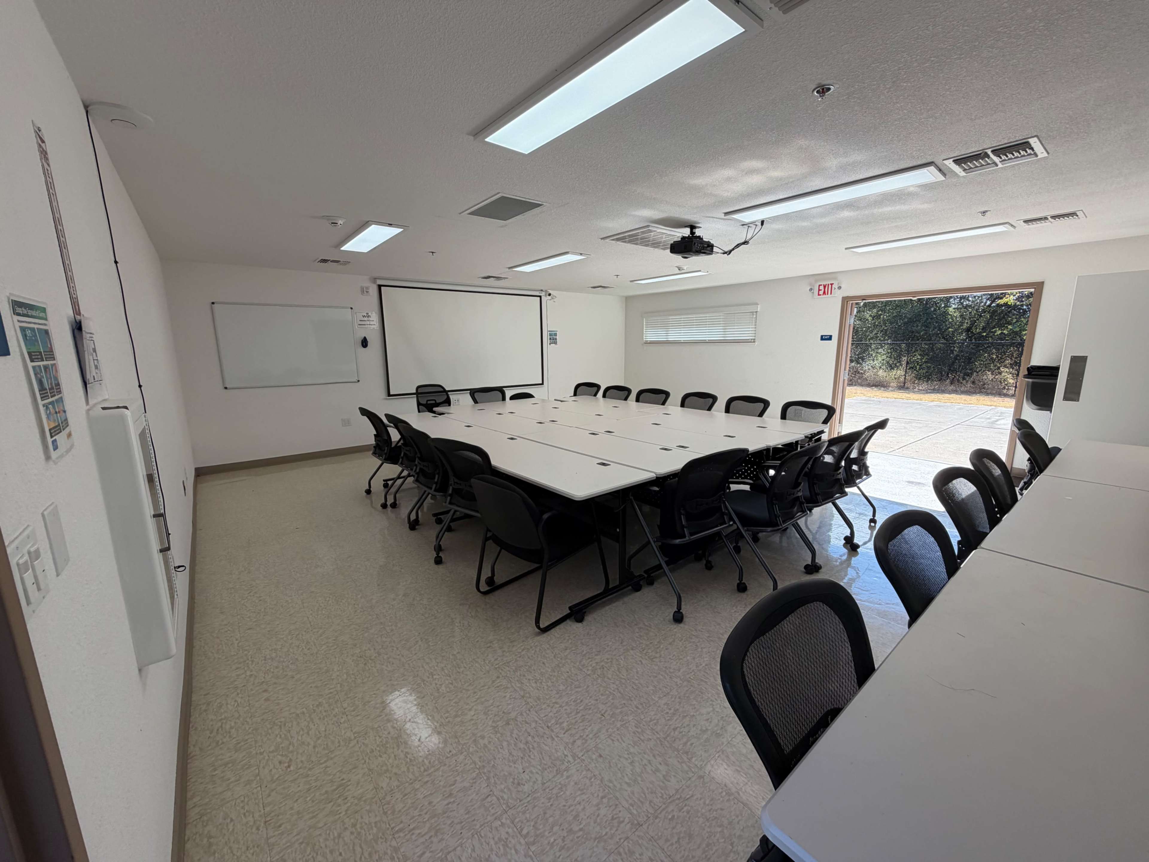 The image shows a conference room equipped with a large rectangular table surrounded by black chairs, whiteboards on the walls, and doors leading outside.