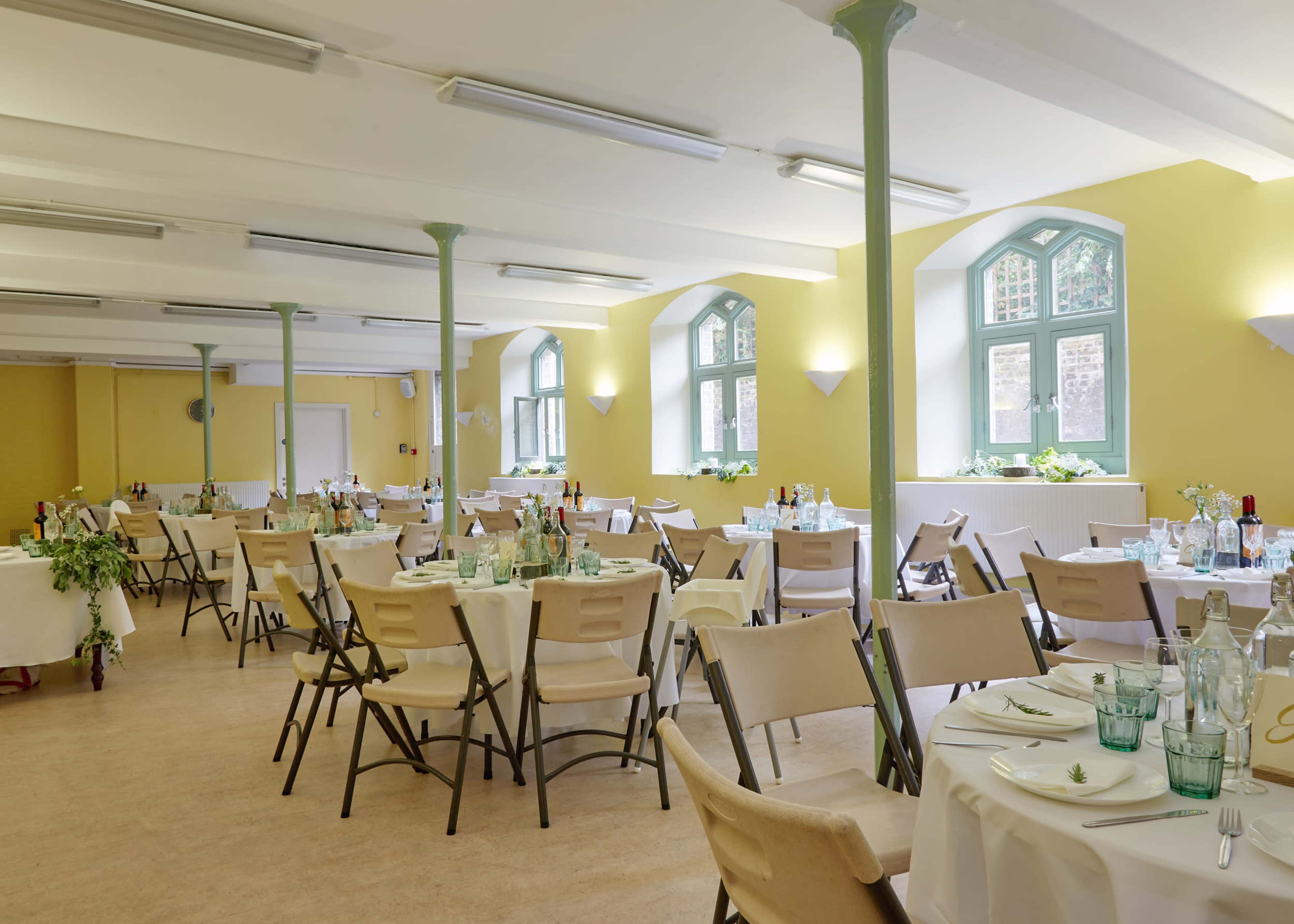 A spacious dining area features numerous tables set for a meal, with green windows and light-colored walls.