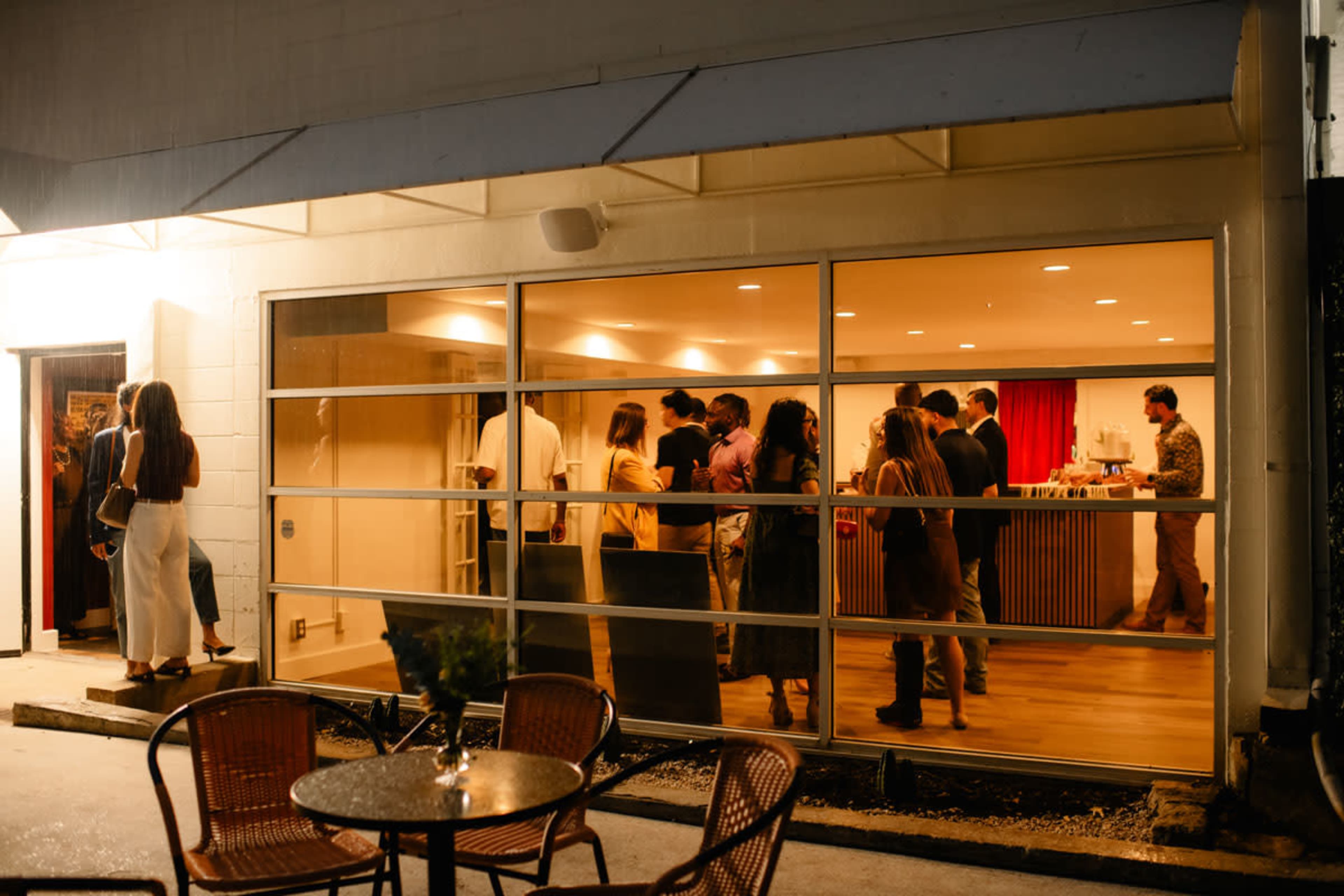 A group of people is gathered inside a brightly lit venue, while a few individuals stand outside near a table with chairs.