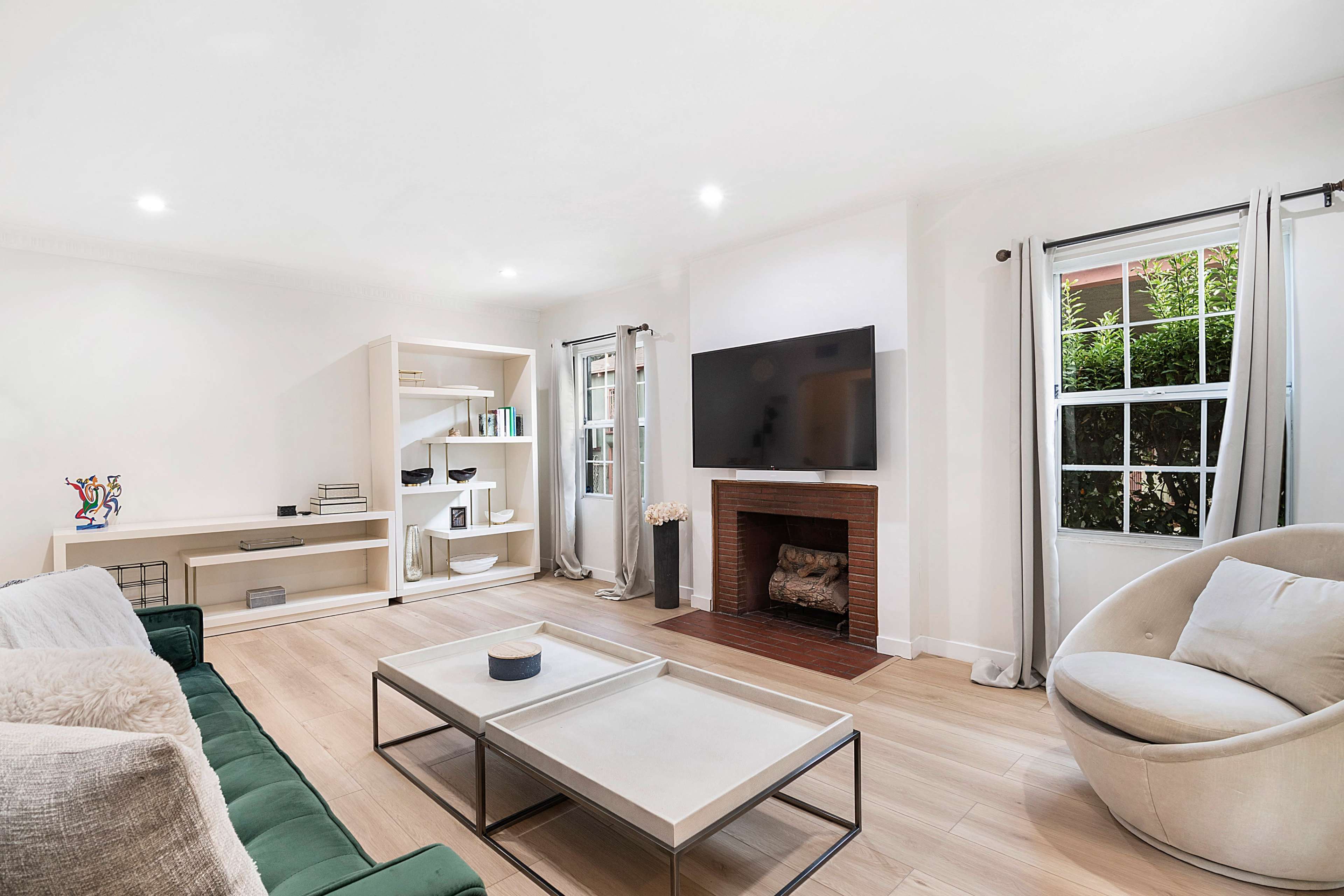 The living room features a green couch, a coffee table, a television mounted on the wall, and large windows with light curtains.