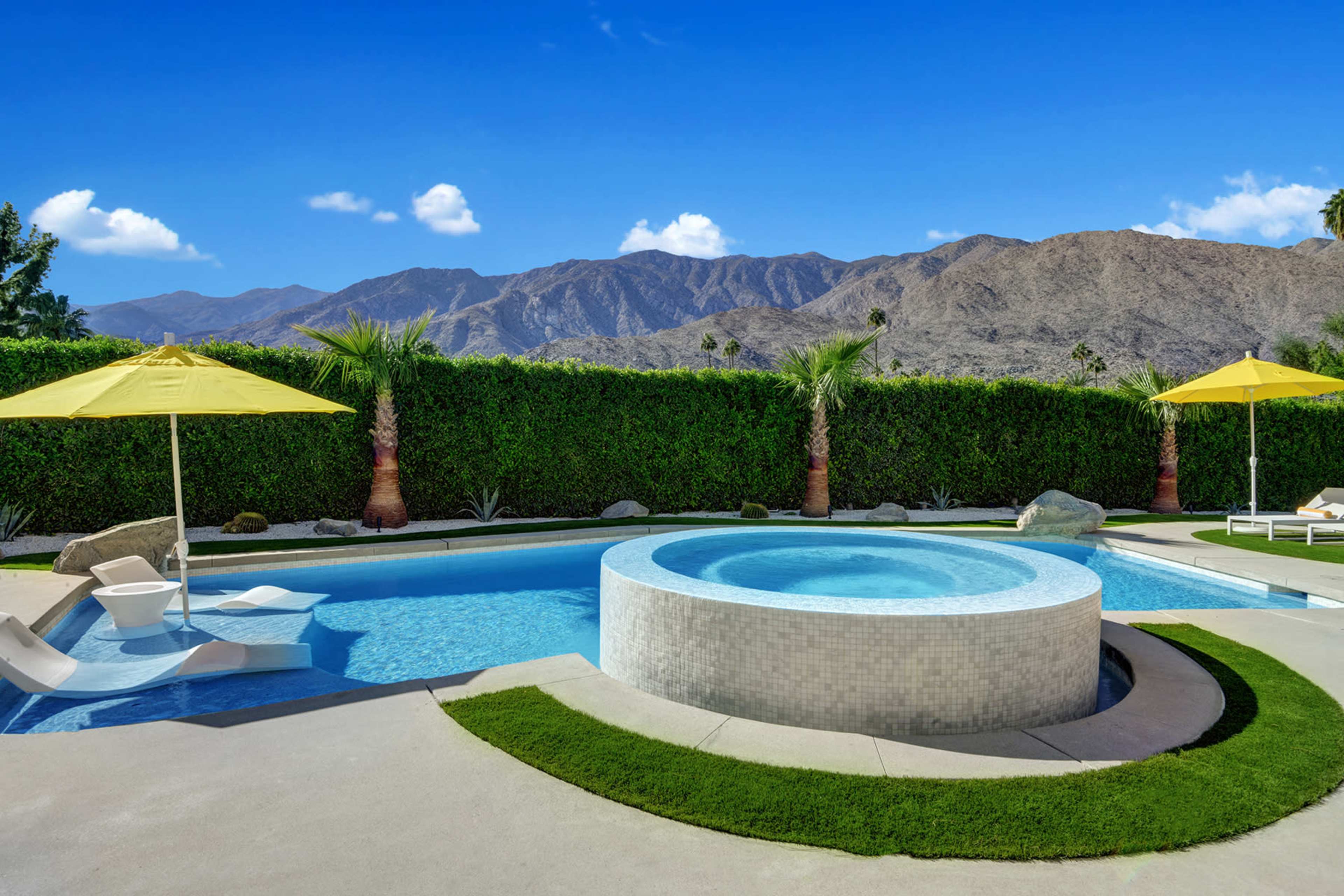 A modern swimming pool with a hot tub, surrounded by green palm trees and a mountain backdrop under a clear blue sky.