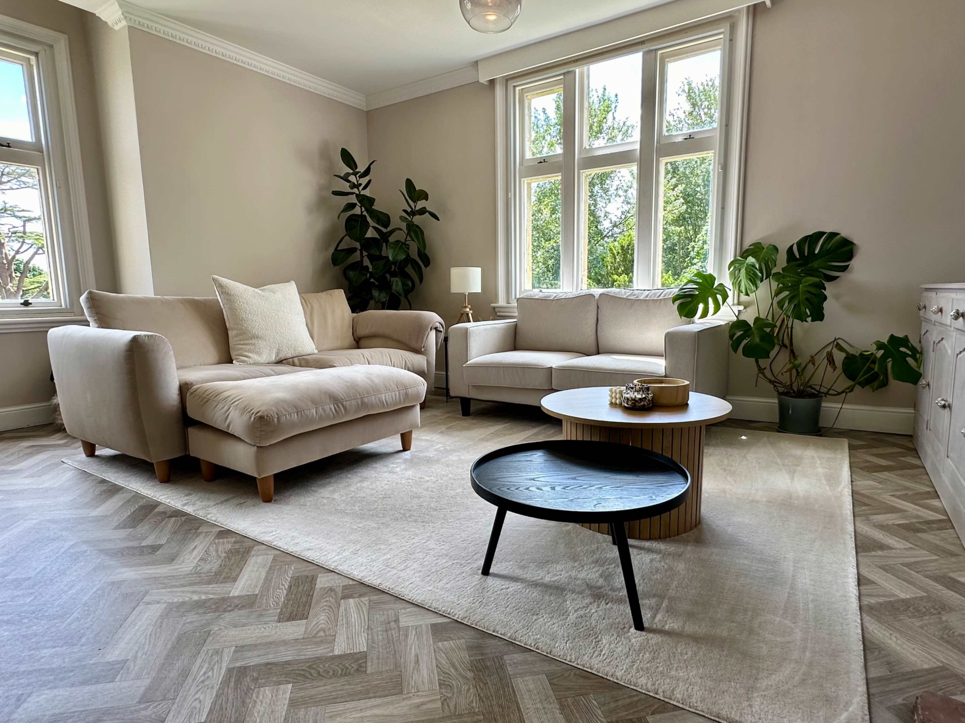 A bright living room features a light-colored sofa and armchair, a round coffee table, and plants by large windows.