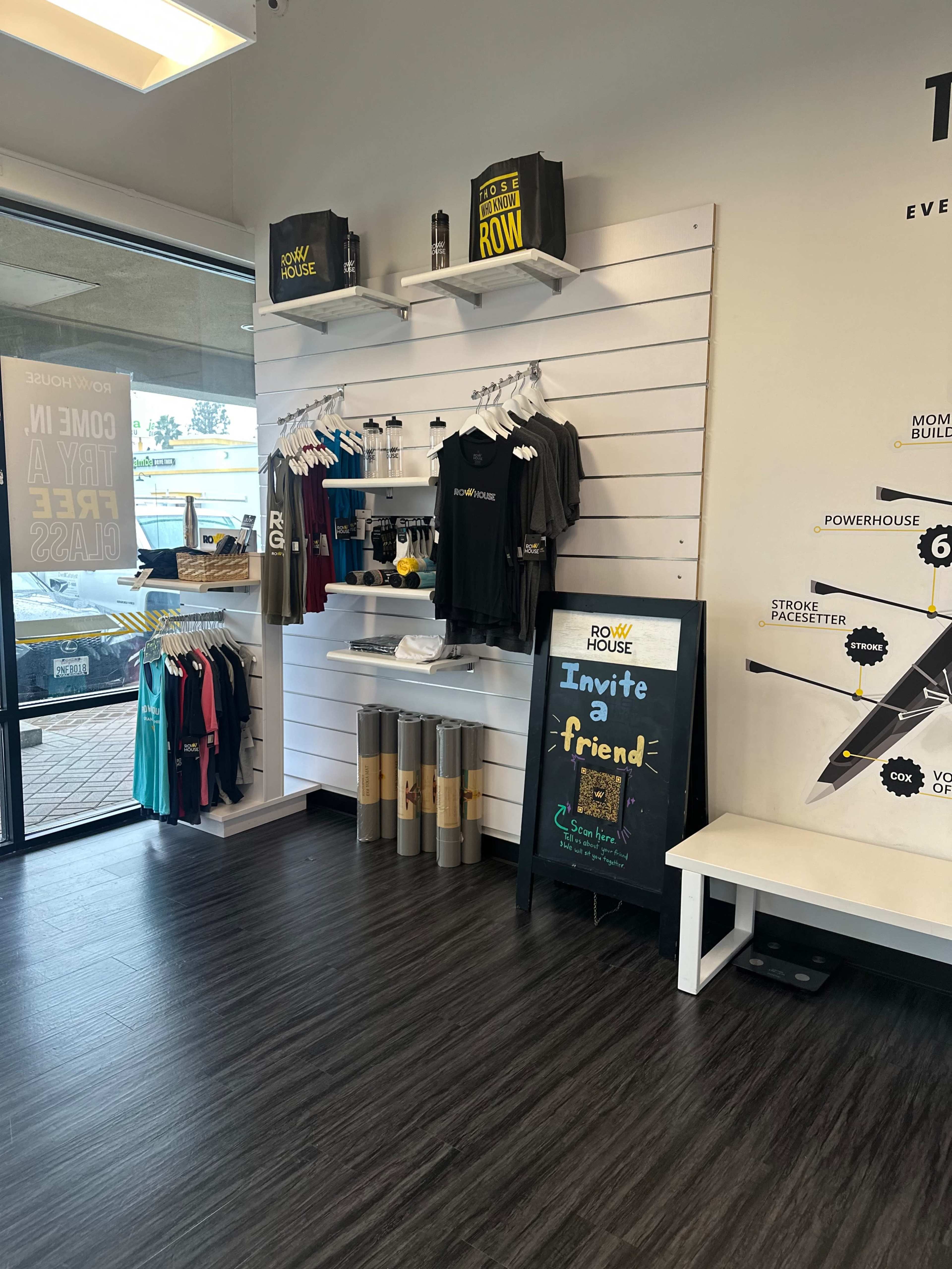 The image shows an interior retail space featuring merchandise such as t-shirts and promotional materials displayed on a wall, with a chalkboard sign inviting people to invite a friend.