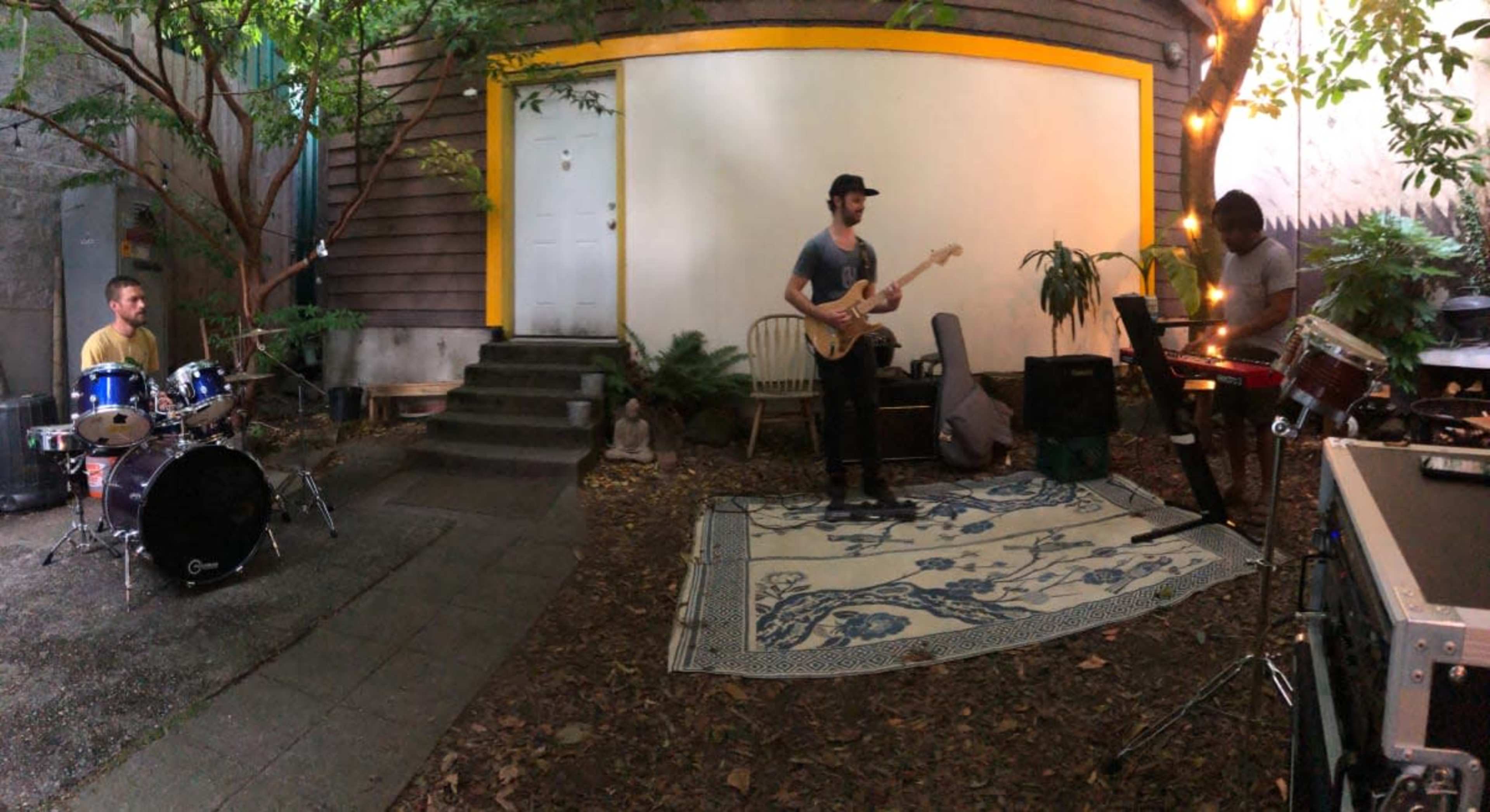 Three musicians perform in a garden space, with one on drums, one on guitar, and one on keyboards, surrounded by plants and decorative lighting.
