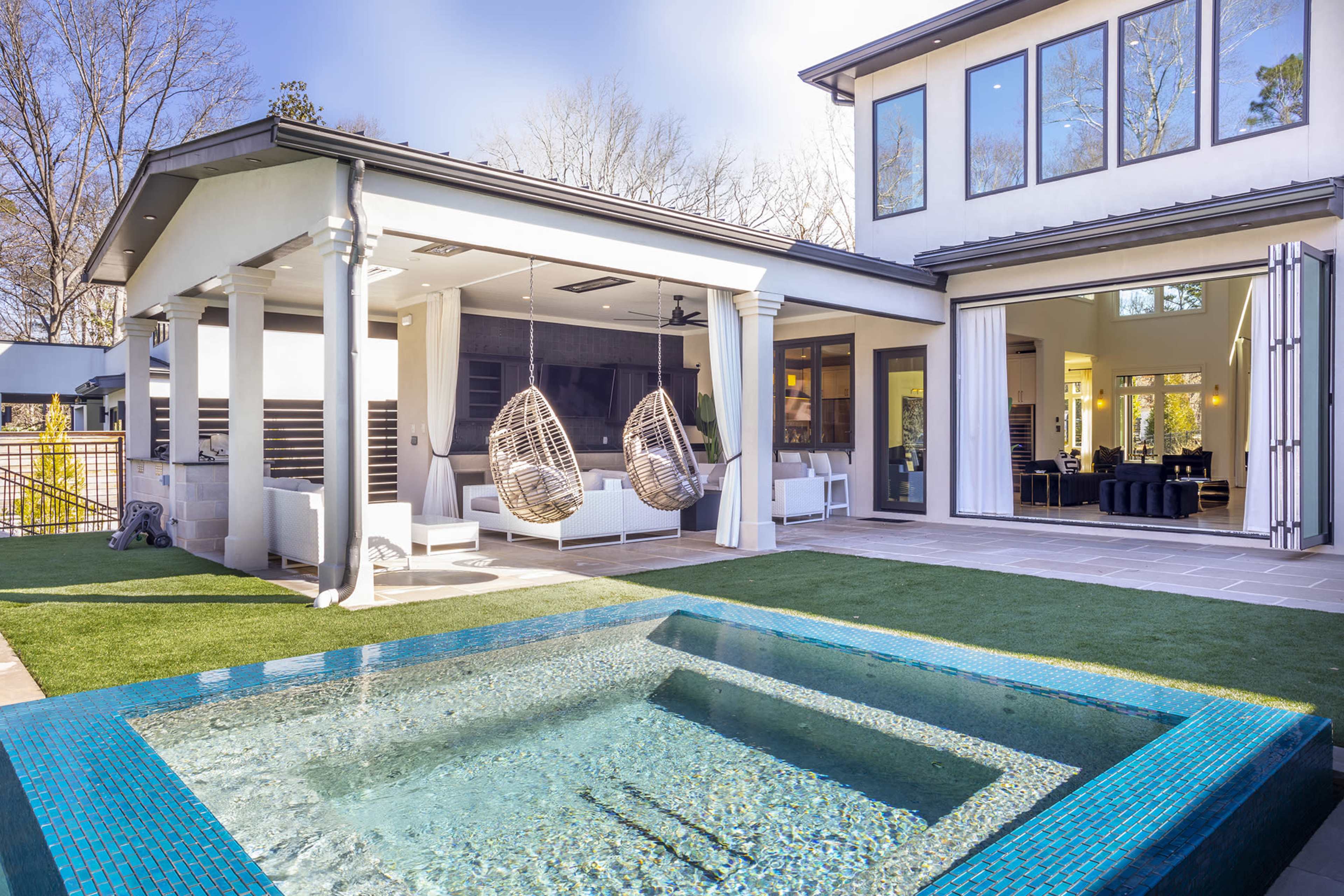 A modern backyard with a small pool, a pair of hanging chairs under a covered area, and a view of a house with large windows and an open interior space.