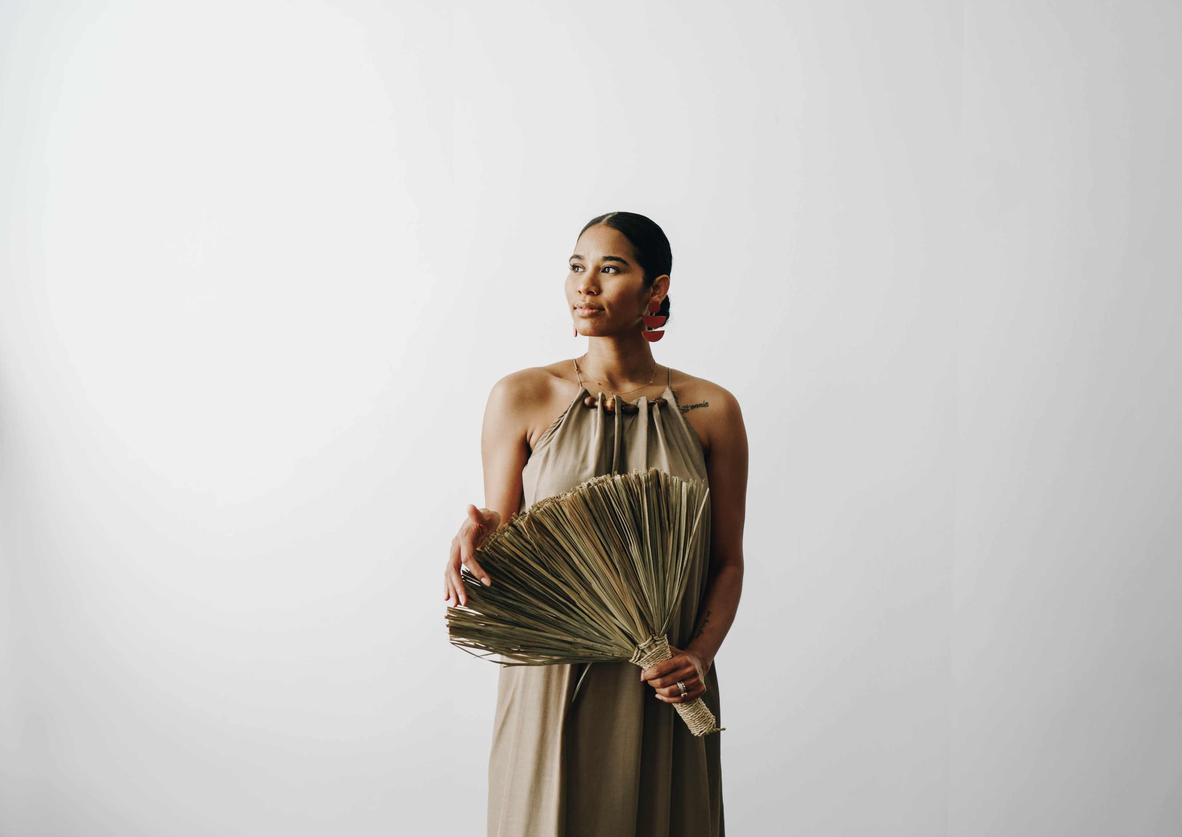A woman stands against a plain white background holding a large, folded fan.