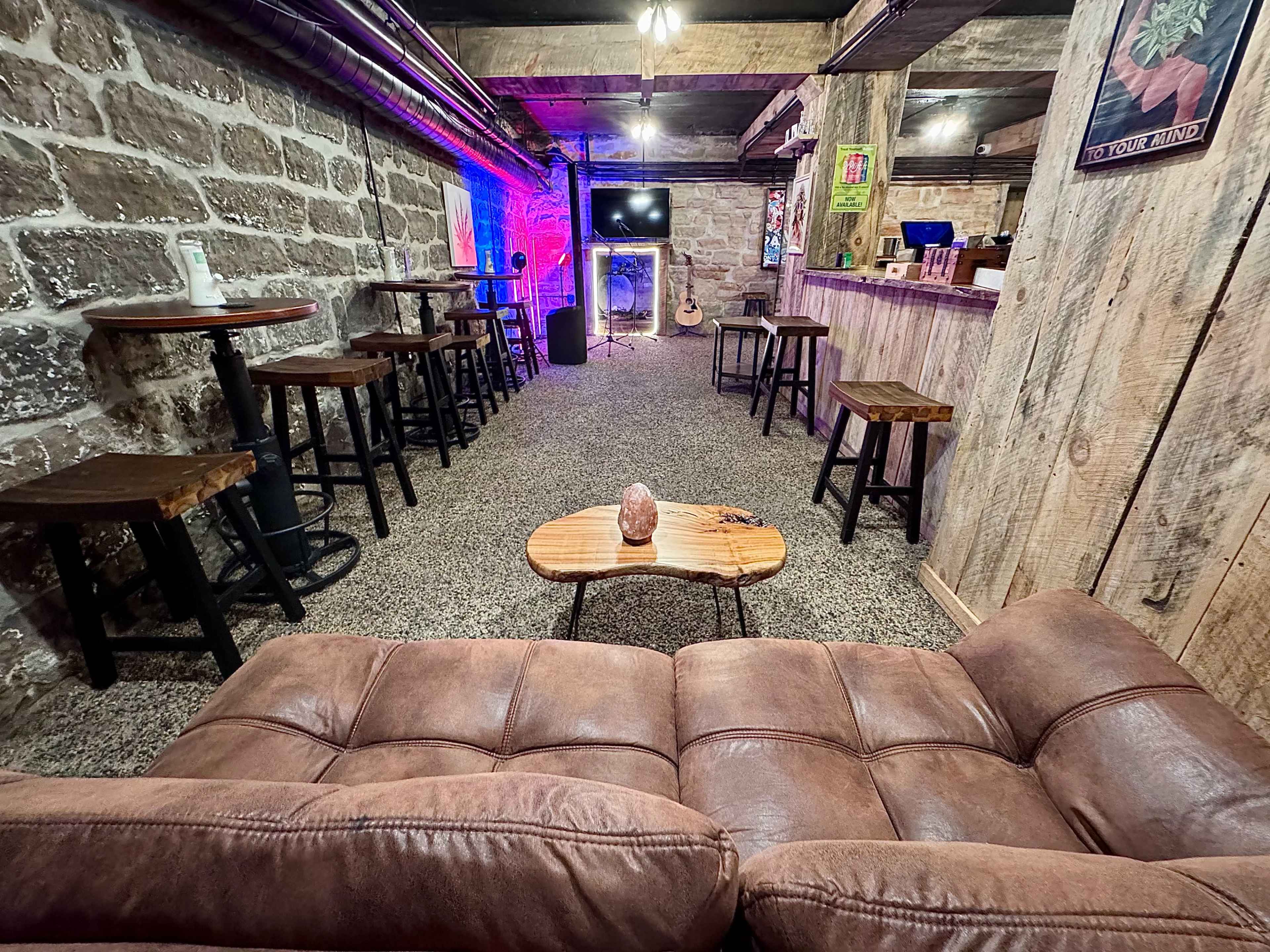The image shows a cozy, rustic lounge area with a brown couch facing several bar stools and a wooden table, illuminated by soft lighting in a stone-walled basement.