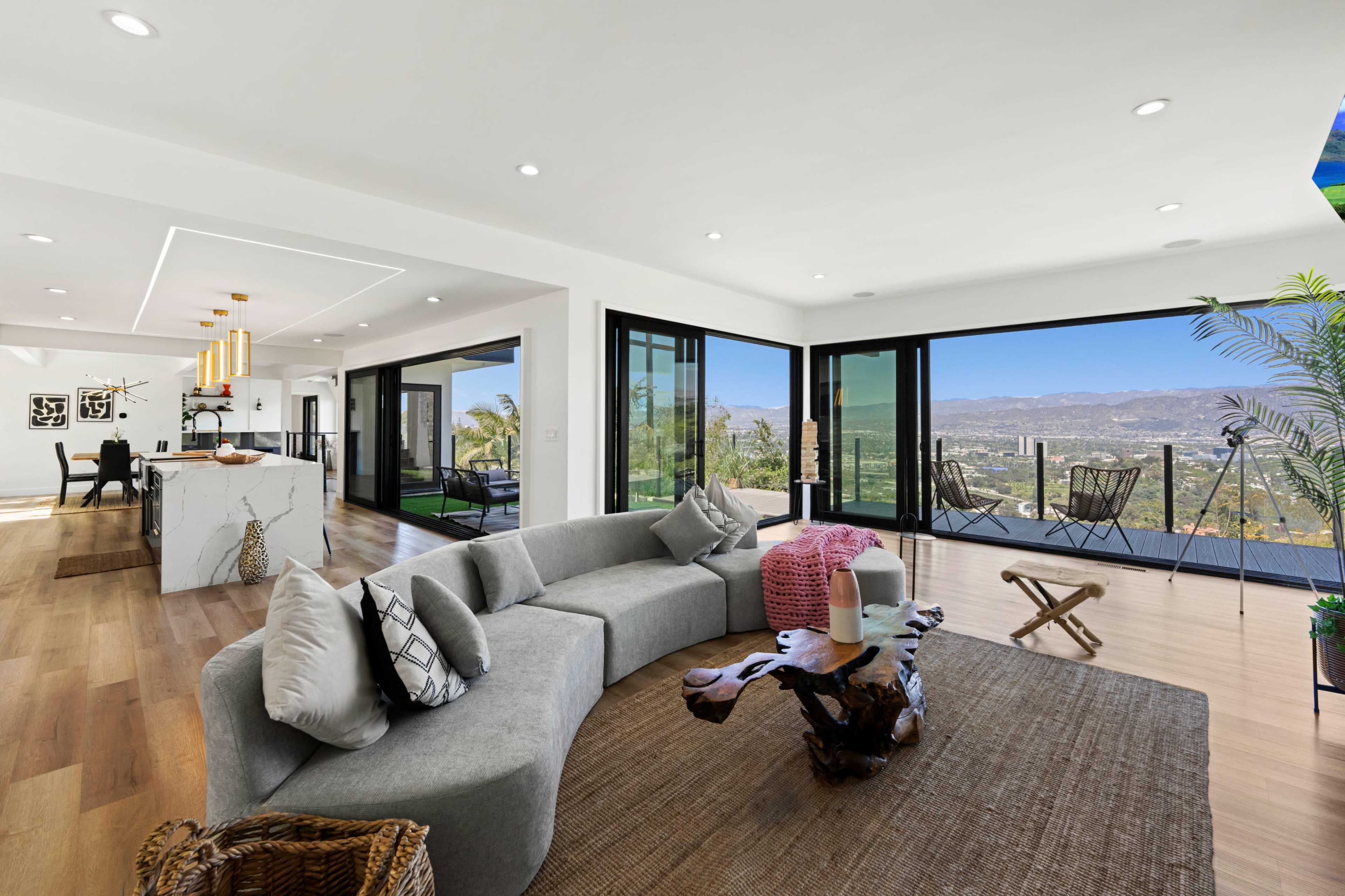 A modern living room featuring a curved gray sofa, a wooden coffee table, and large glass sliding doors that open to a mountain view.