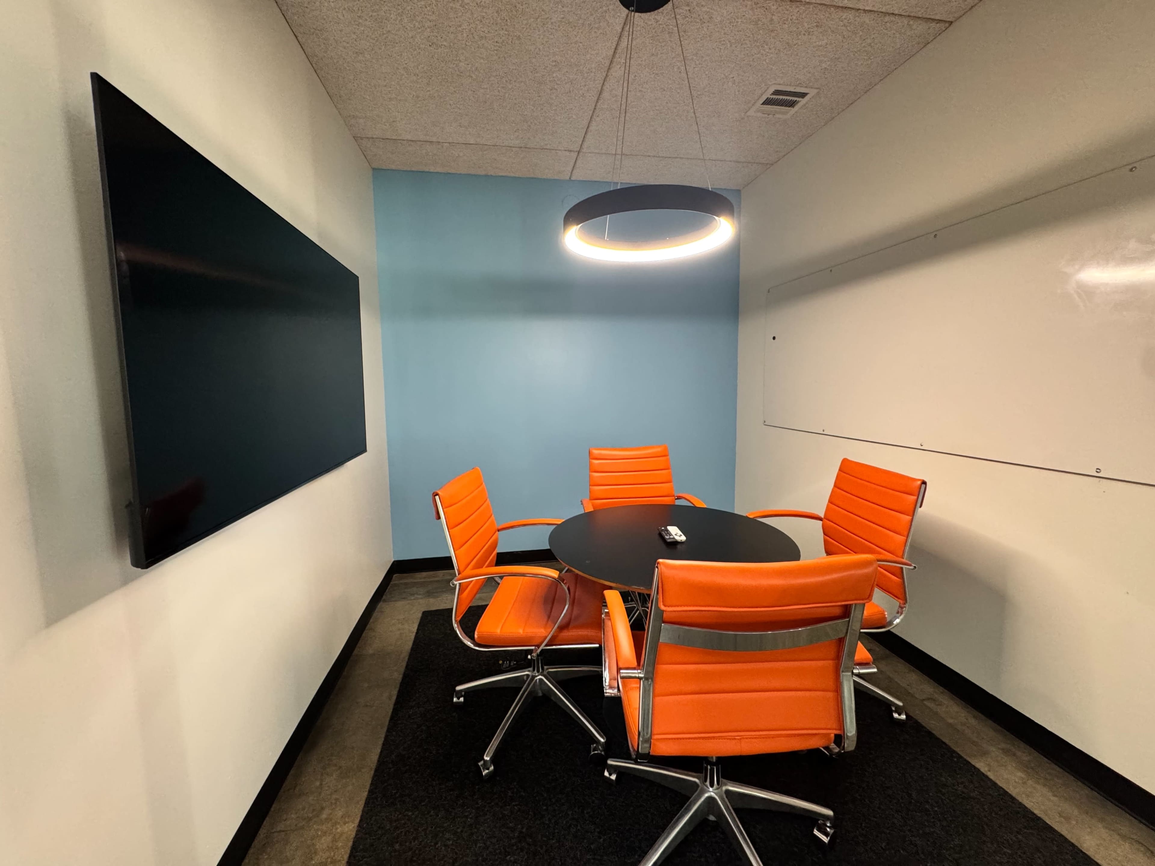 A small meeting room features a round black table surrounded by four orange chairs, with a large screen on one wall and a whiteboard on another.