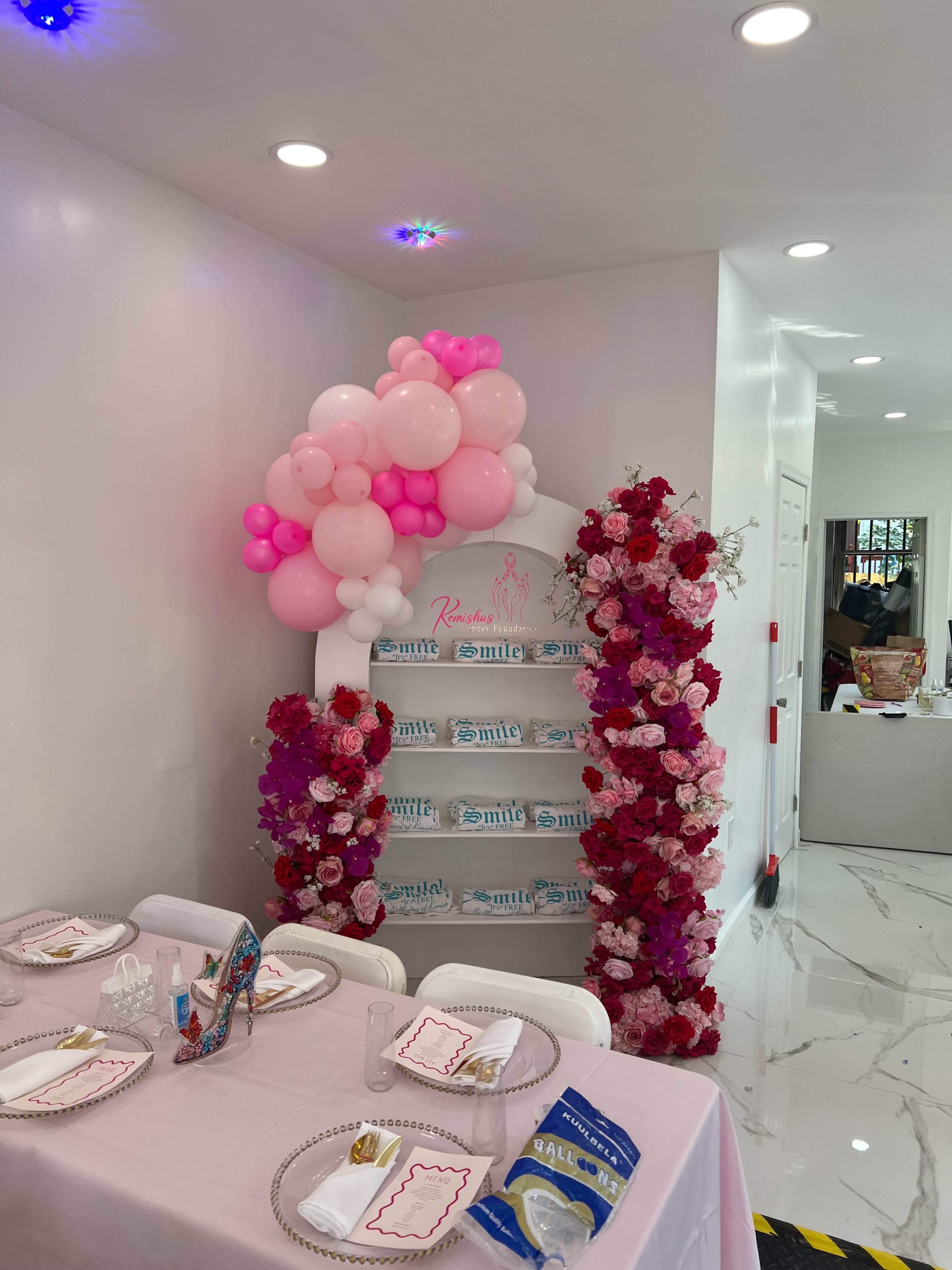 The image shows a decorated corner of a room with a balloon arch in shades of pink, surrounded by floral arrangements, and a table set for an event featuring elegant tableware.