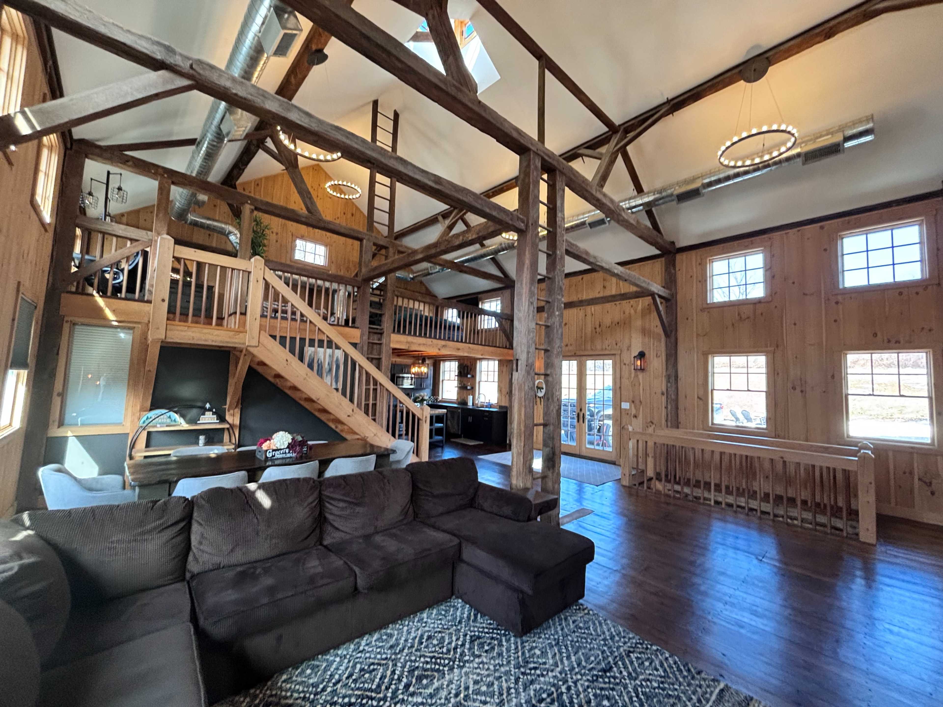 The image shows a spacious, rustic interior of a wooden lodge featuring a living area with a large sectional sofa, a dining table, and a visible staircase leading to a loft area.