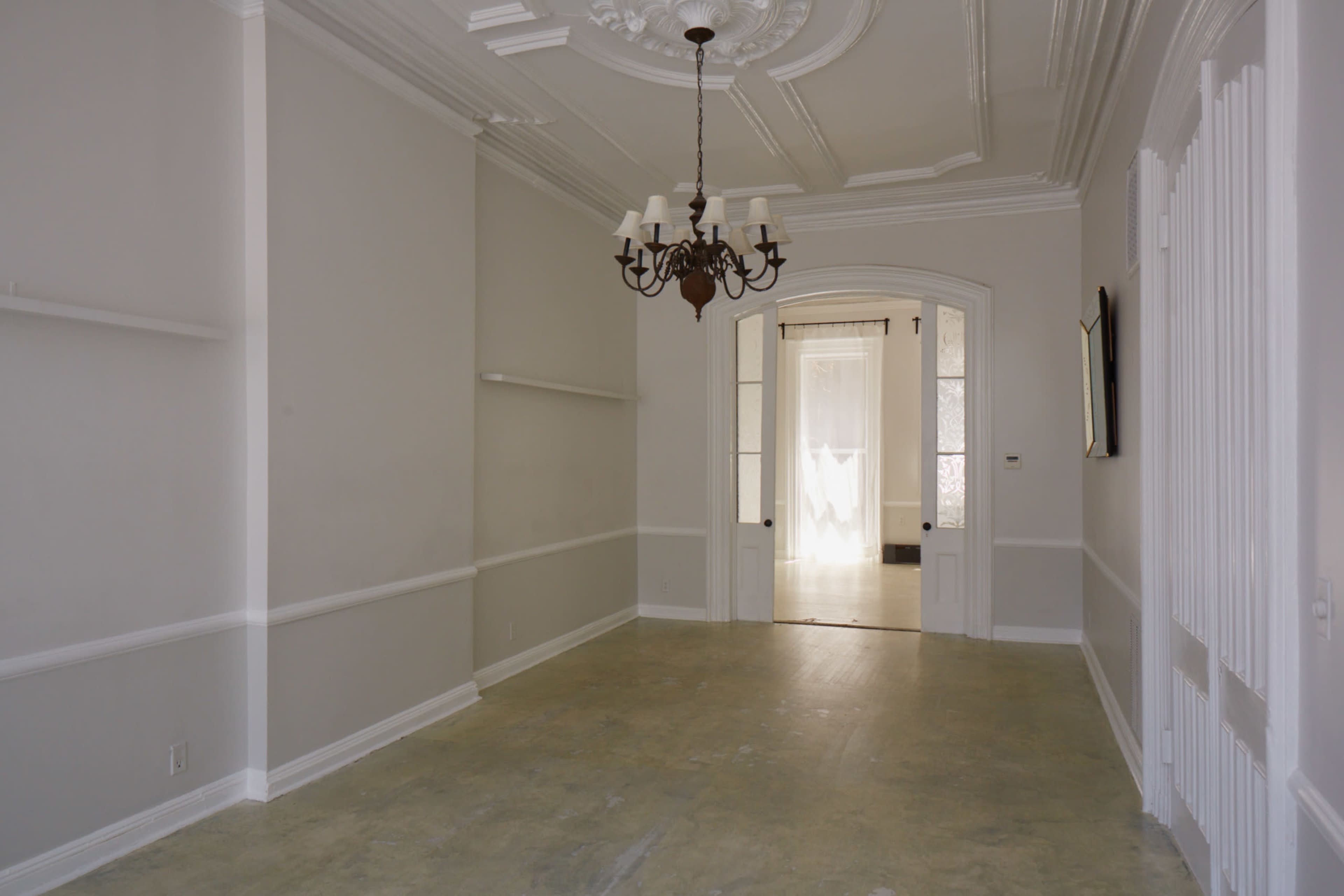 A spacious room features a chandelier, ornate ceiling details, and arched doorways leading to other areas.