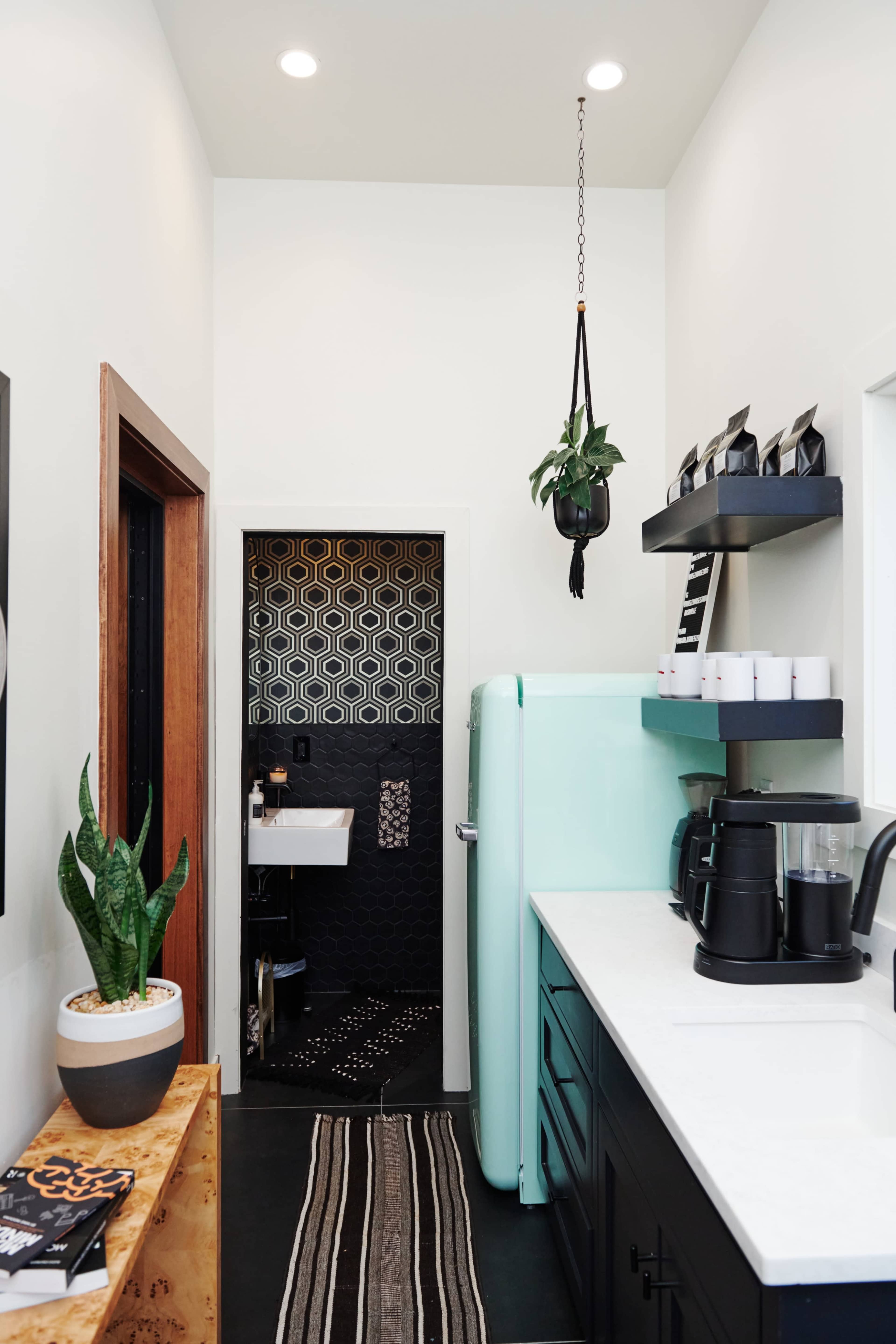 The image shows a small, modern kitchenette featuring a mint green refrigerator, black cabinetry, and a hanging plant, with a patterned backsplash in the adjacent space.