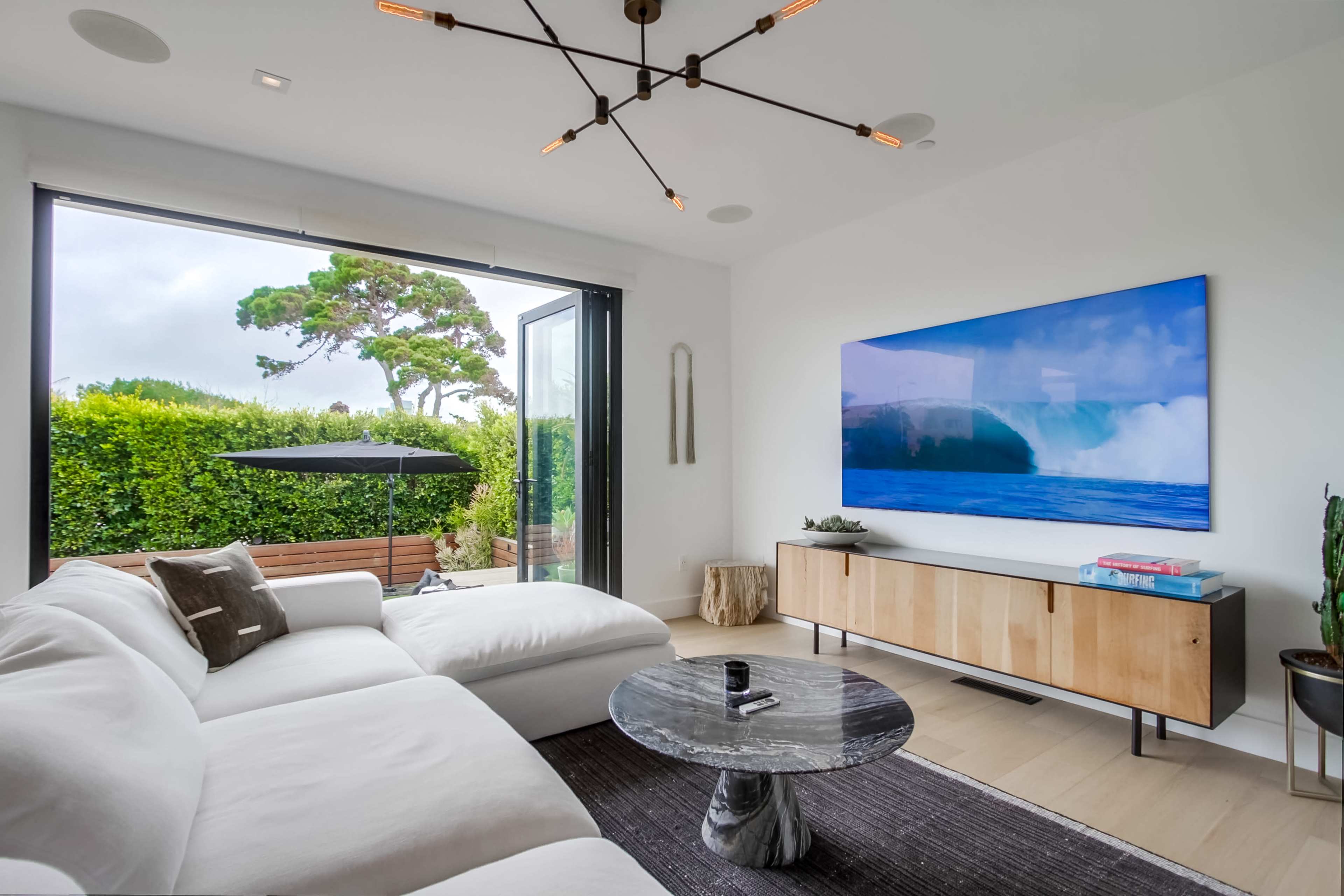 A modern living room features a large white sofa, a round black marble coffee table, and a television mounted on a light wood console, with large glass doors opening to a patio with greenery outside.