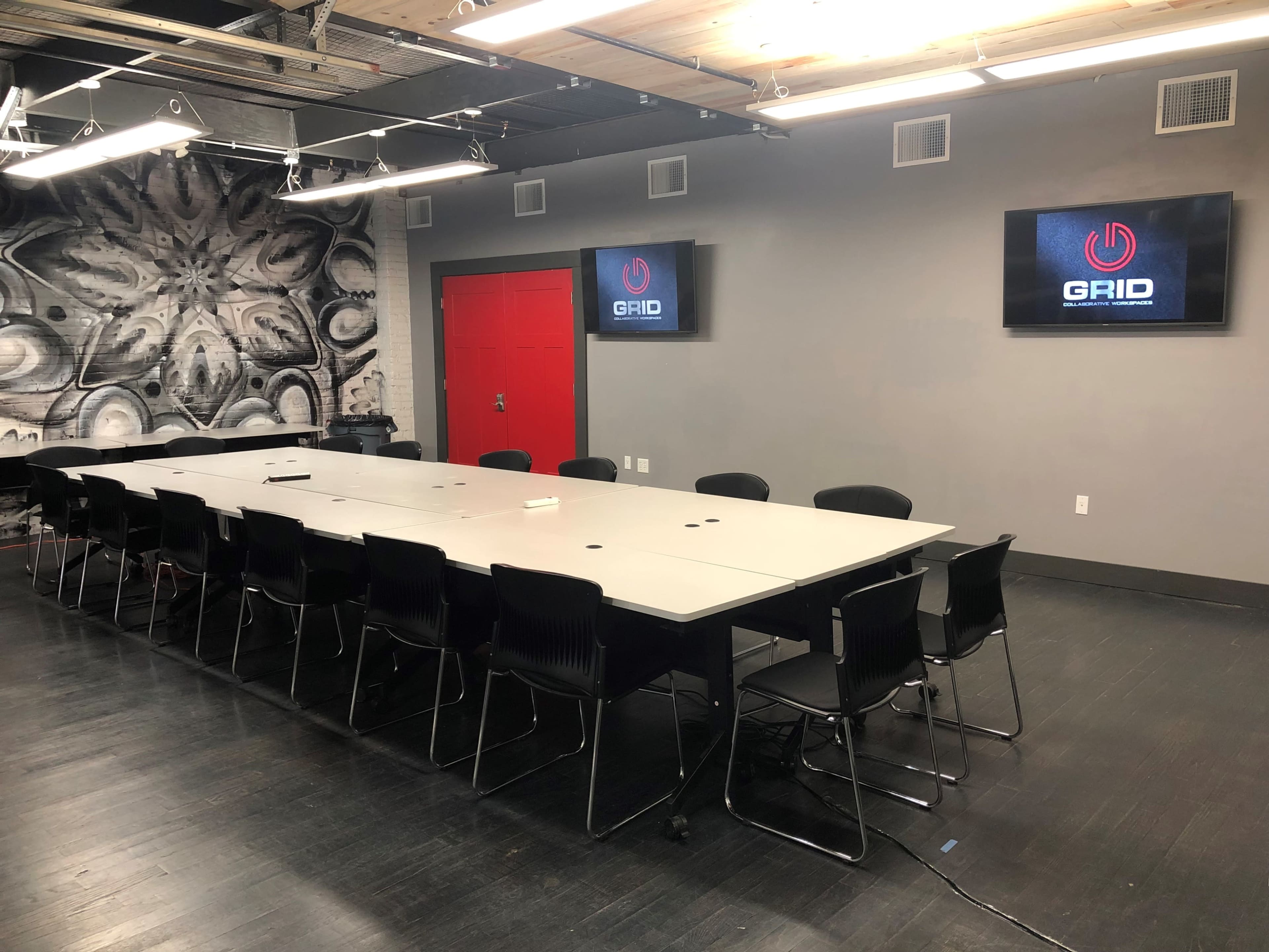 The image shows a conference room with a long table surrounded by black chairs, featuring two screens displaying the logo of "GRID" and a large mural on one wall.