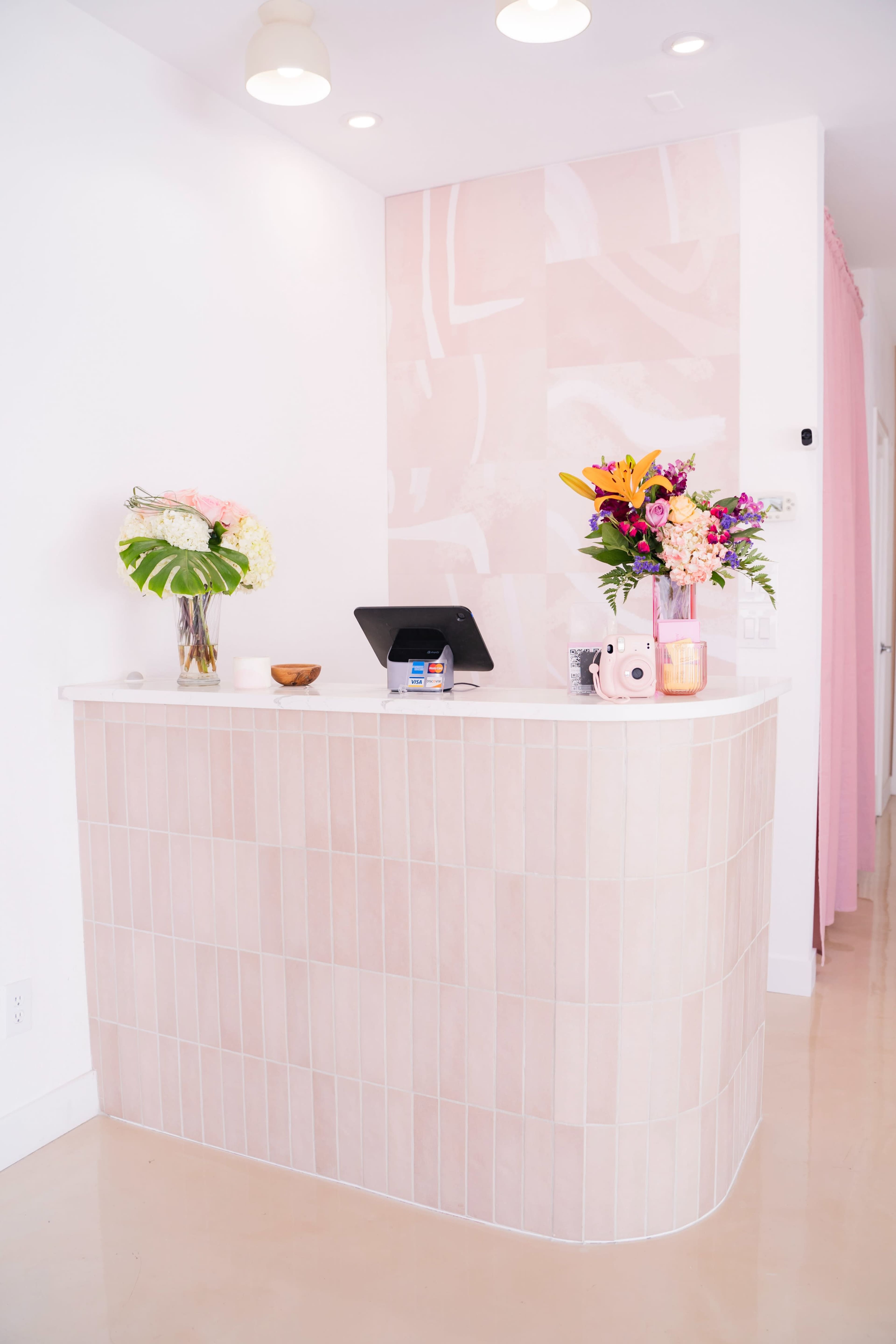 A modern reception desk made of pink tile, complemented by floral arrangements on the surface and a tablet for check-in.