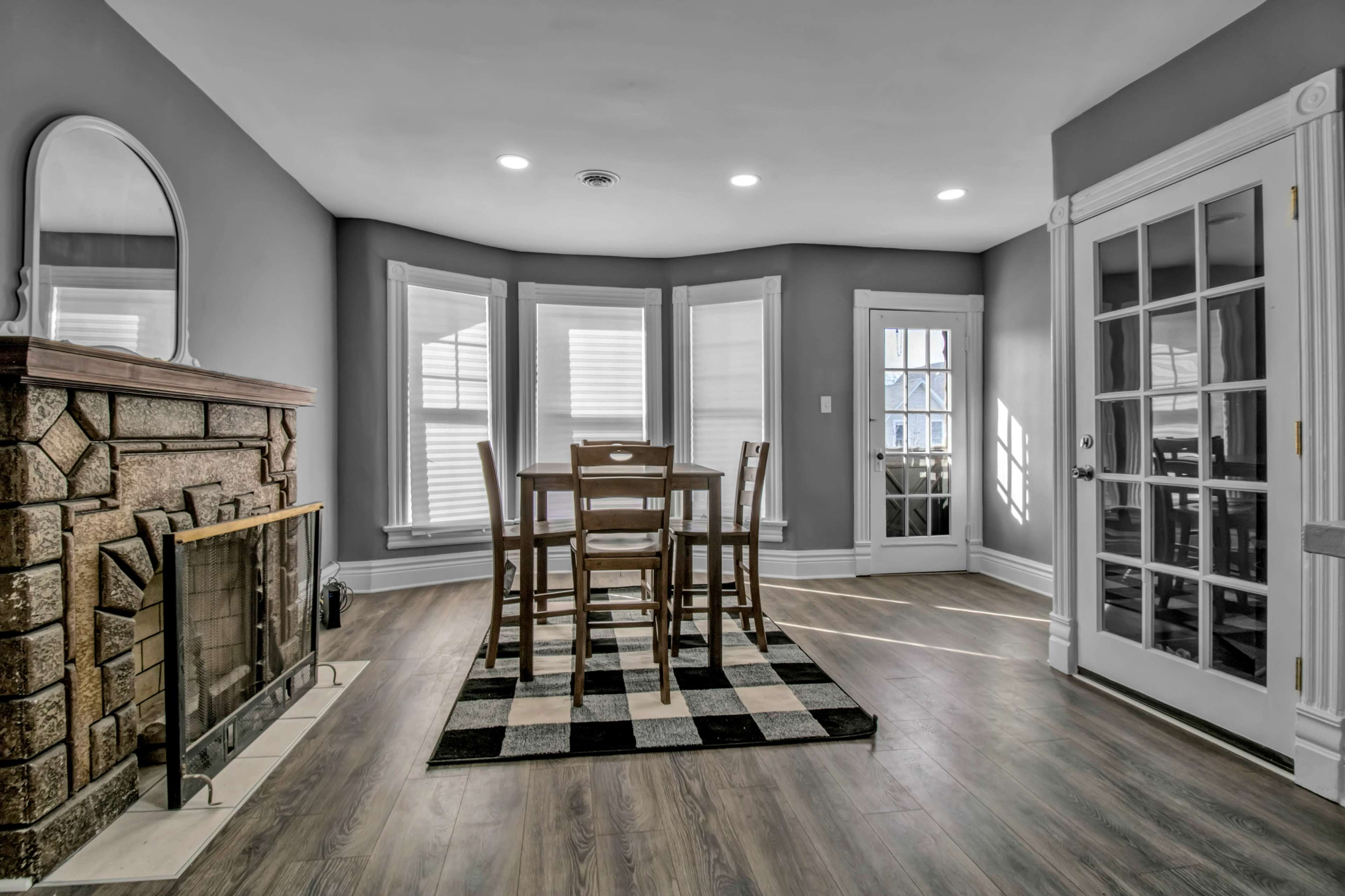 A dining area with a wooden table and chairs, a stone fireplace, and a black-and-white checkered rug, illuminated by natural light from multiple windows.