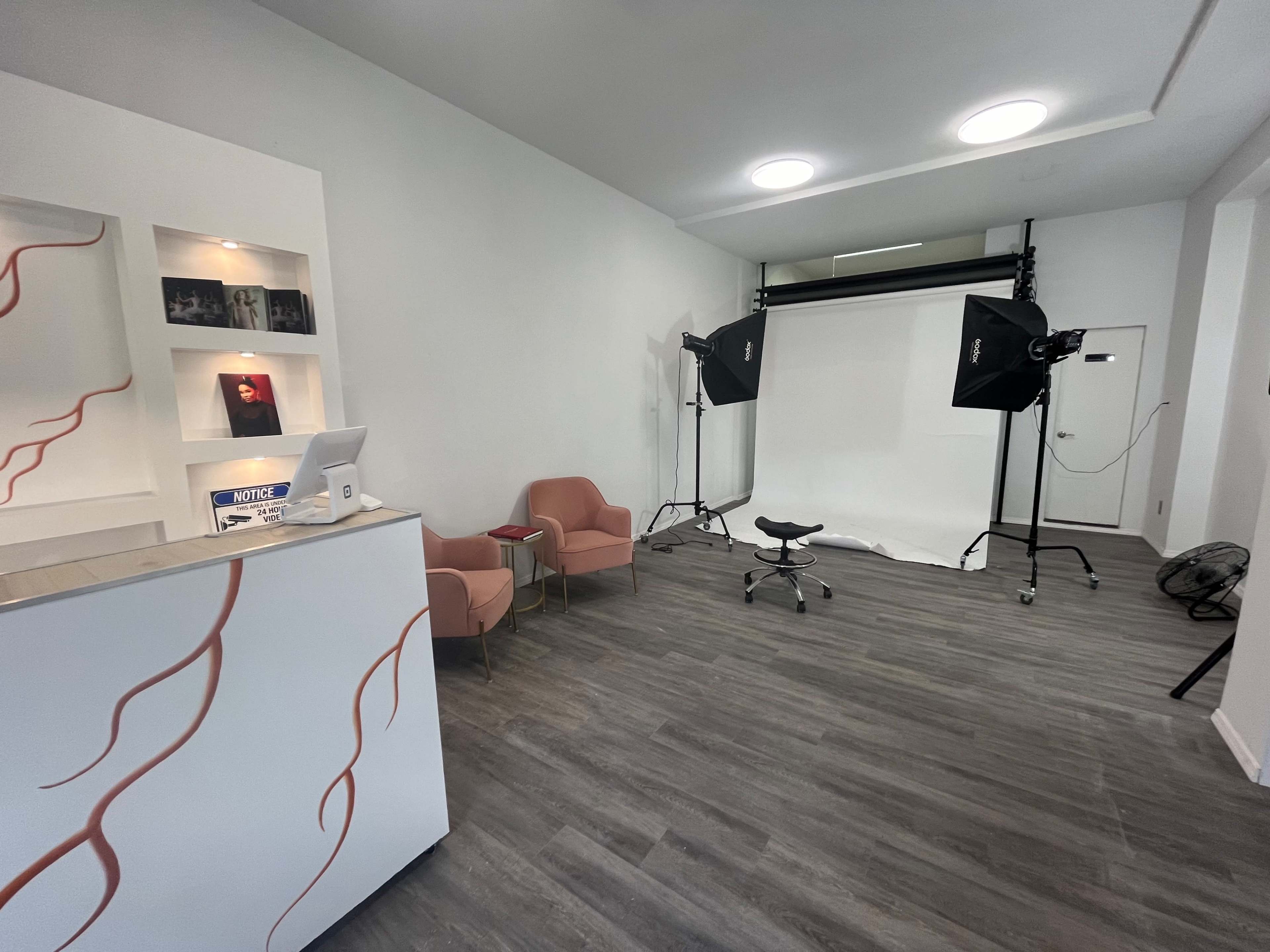 A modern photo studio with a reception area featuring two pink chairs and photography equipment set up against a white backdrop.