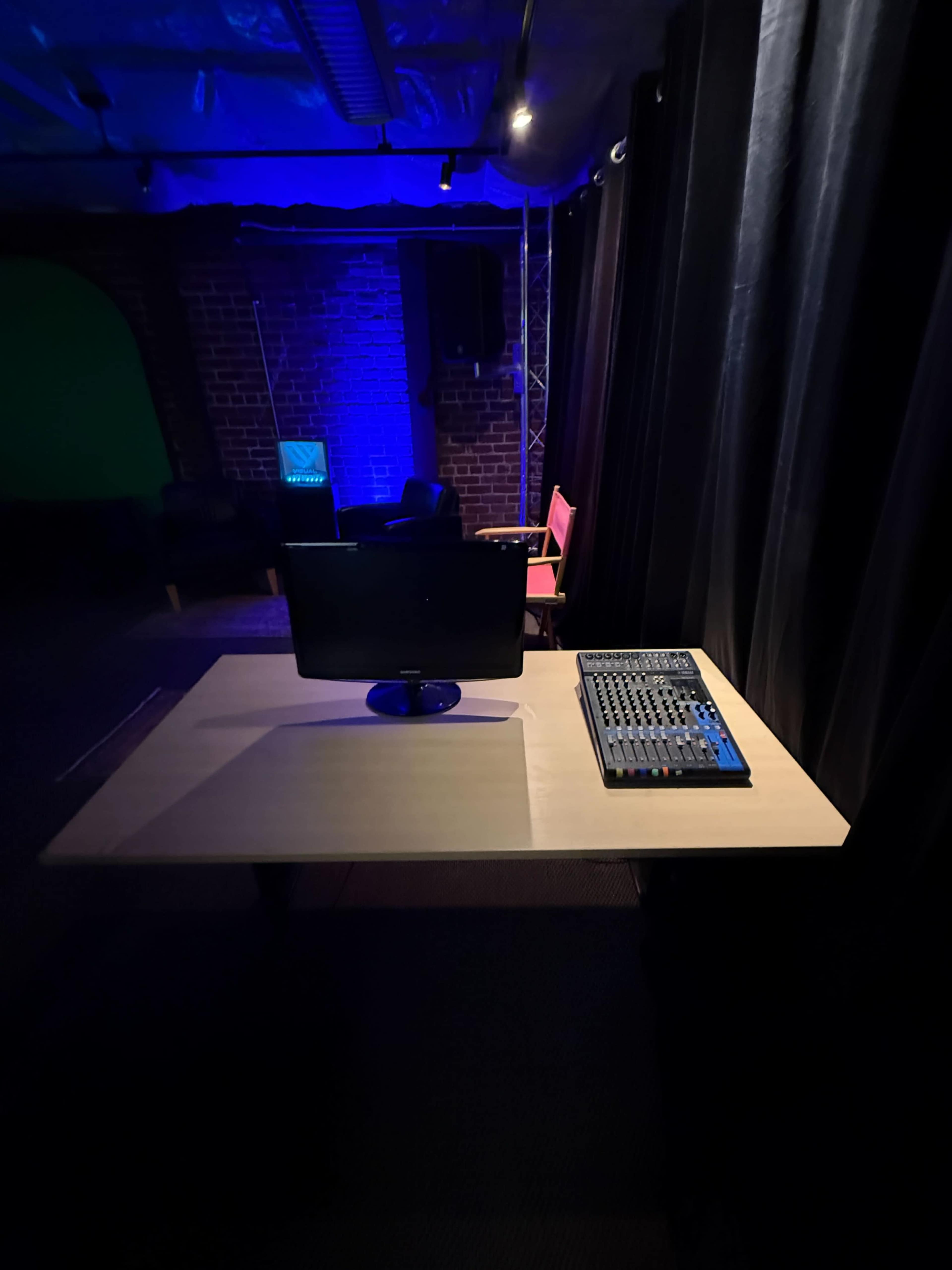 The image shows a workspace with a monitor and a mixing console on a table, surrounded by dark curtains and backdrop lighting.