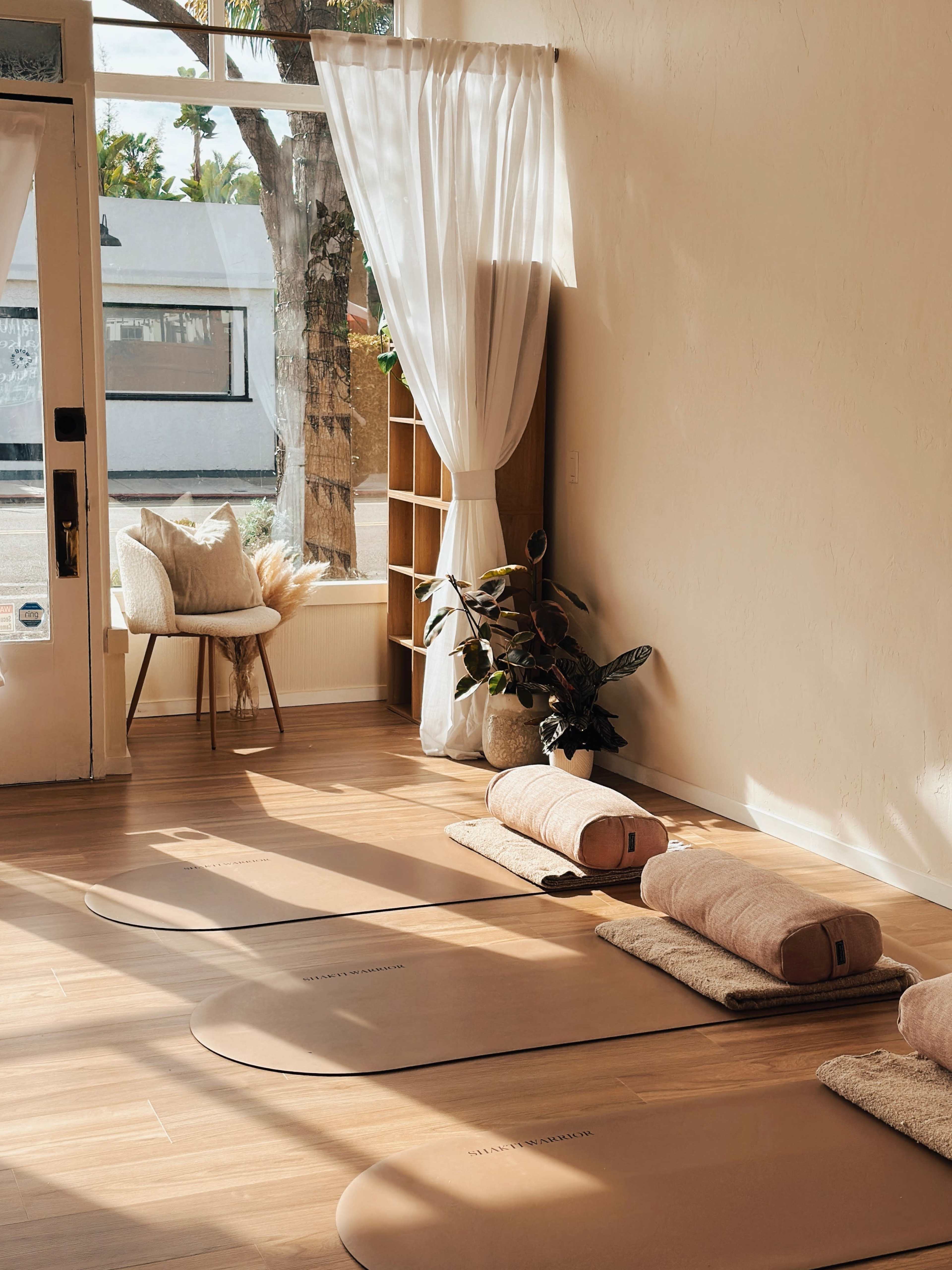 The image shows a serene yoga studio with three mats laid out, soft cushions, and natural light streaming through a large window.