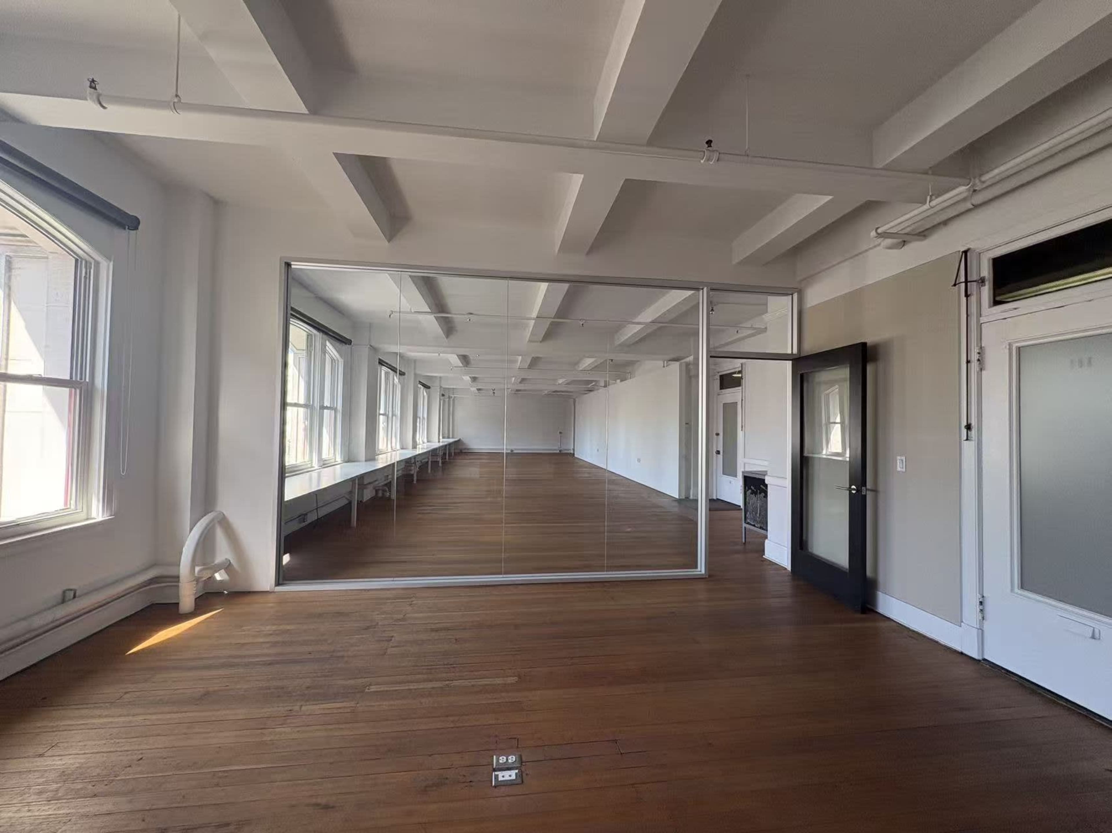 The image shows an empty room with wooden flooring, large windows, and a mirrored wall on one side.