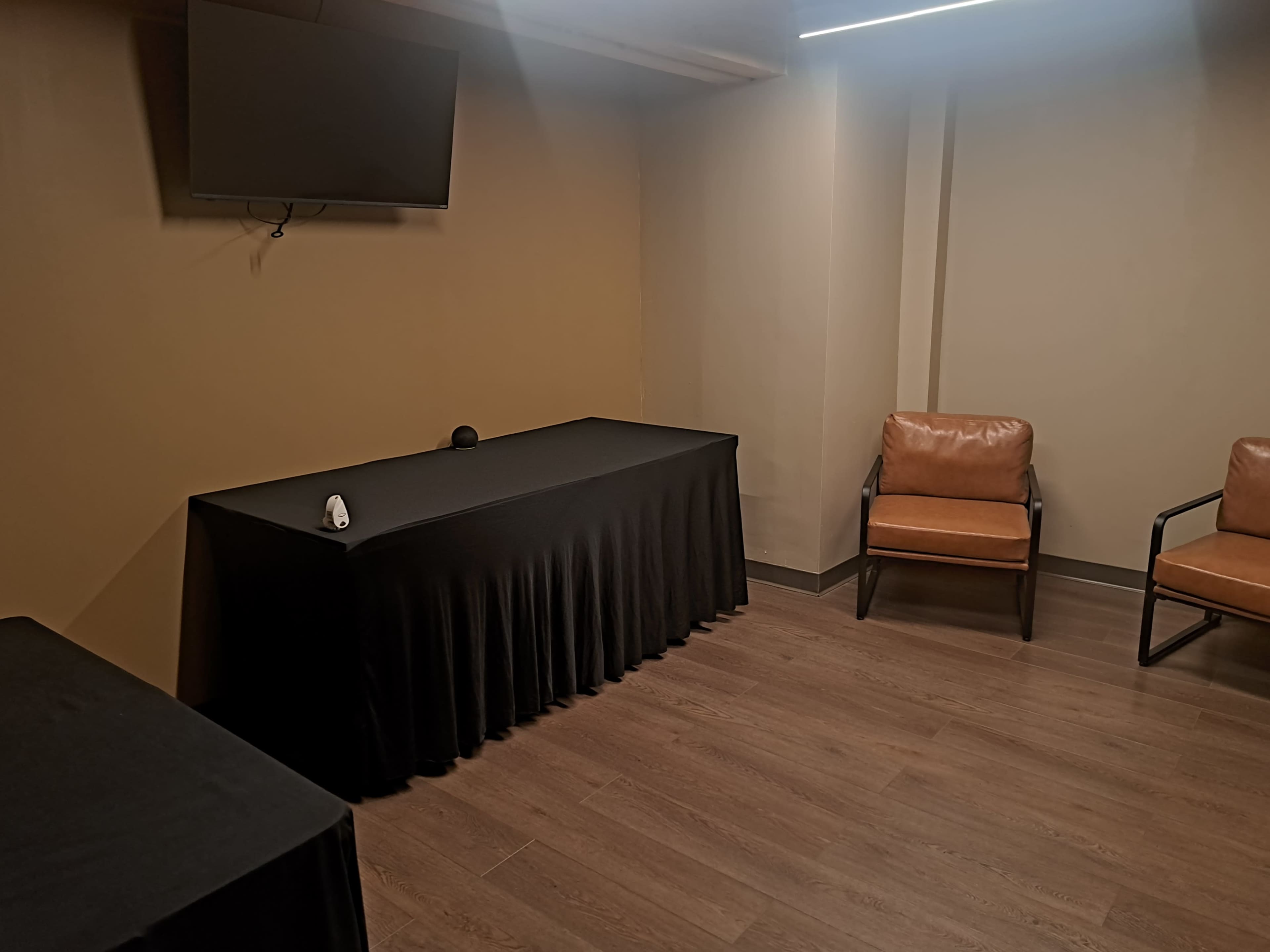 The image shows a small, empty room with a television mounted on the wall, a table covered with a black tablecloth, and two leather chairs.