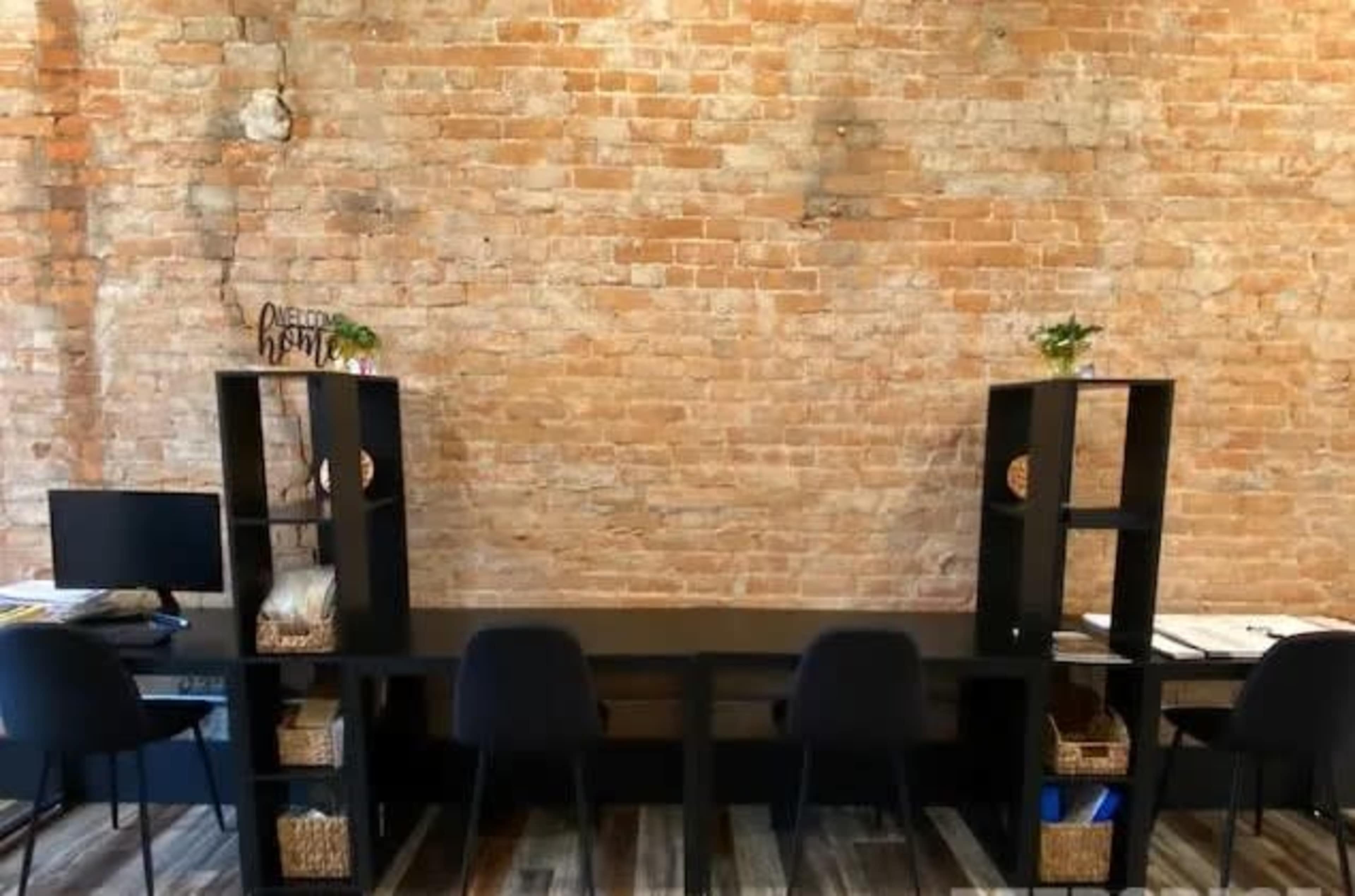 A workspace featuring a long table with black chairs, flanked by two black shelving units against a rustic brick wall.