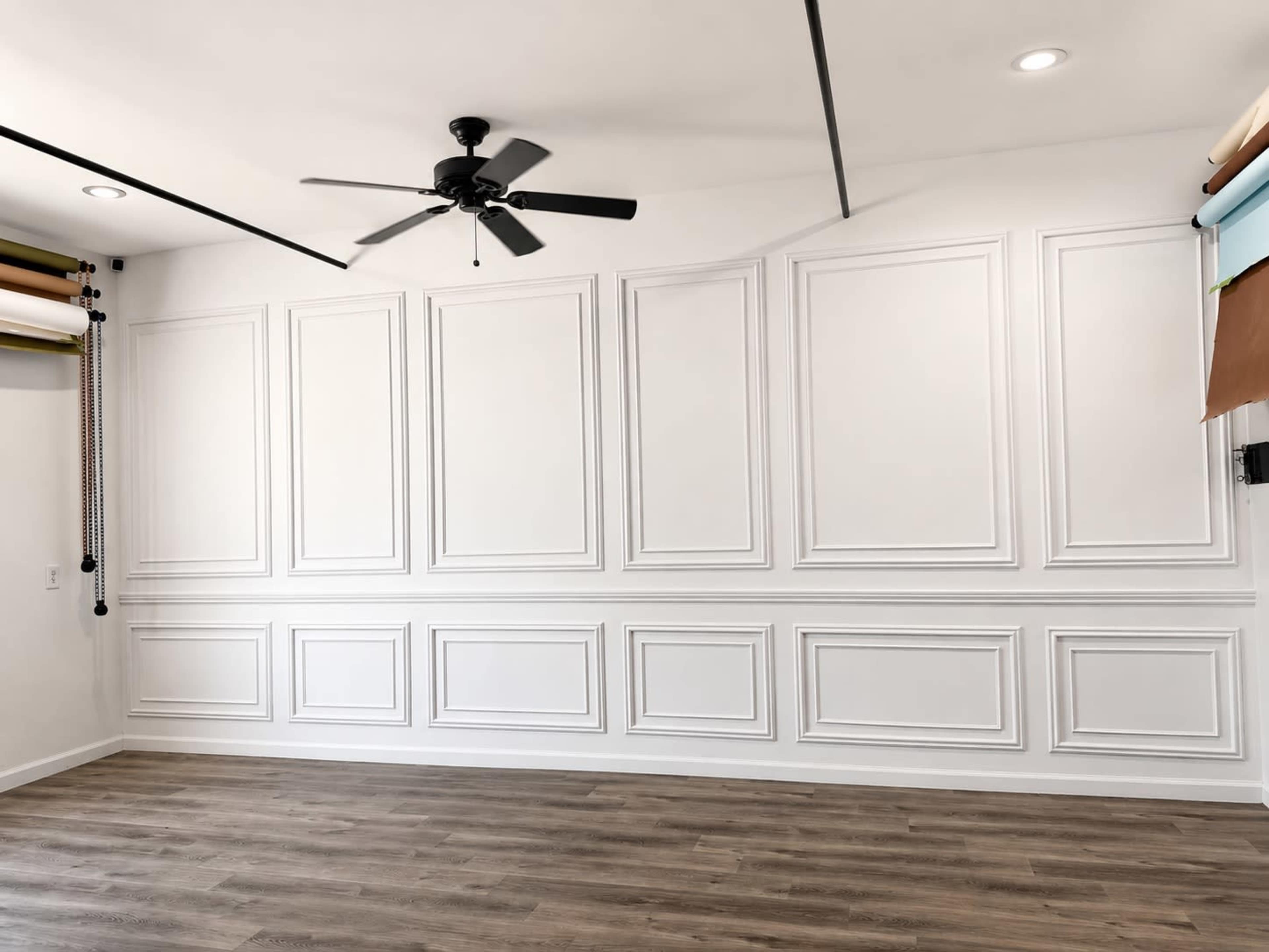 The image shows a spacious, well-lit room with a white paneled wall and a ceiling fan.