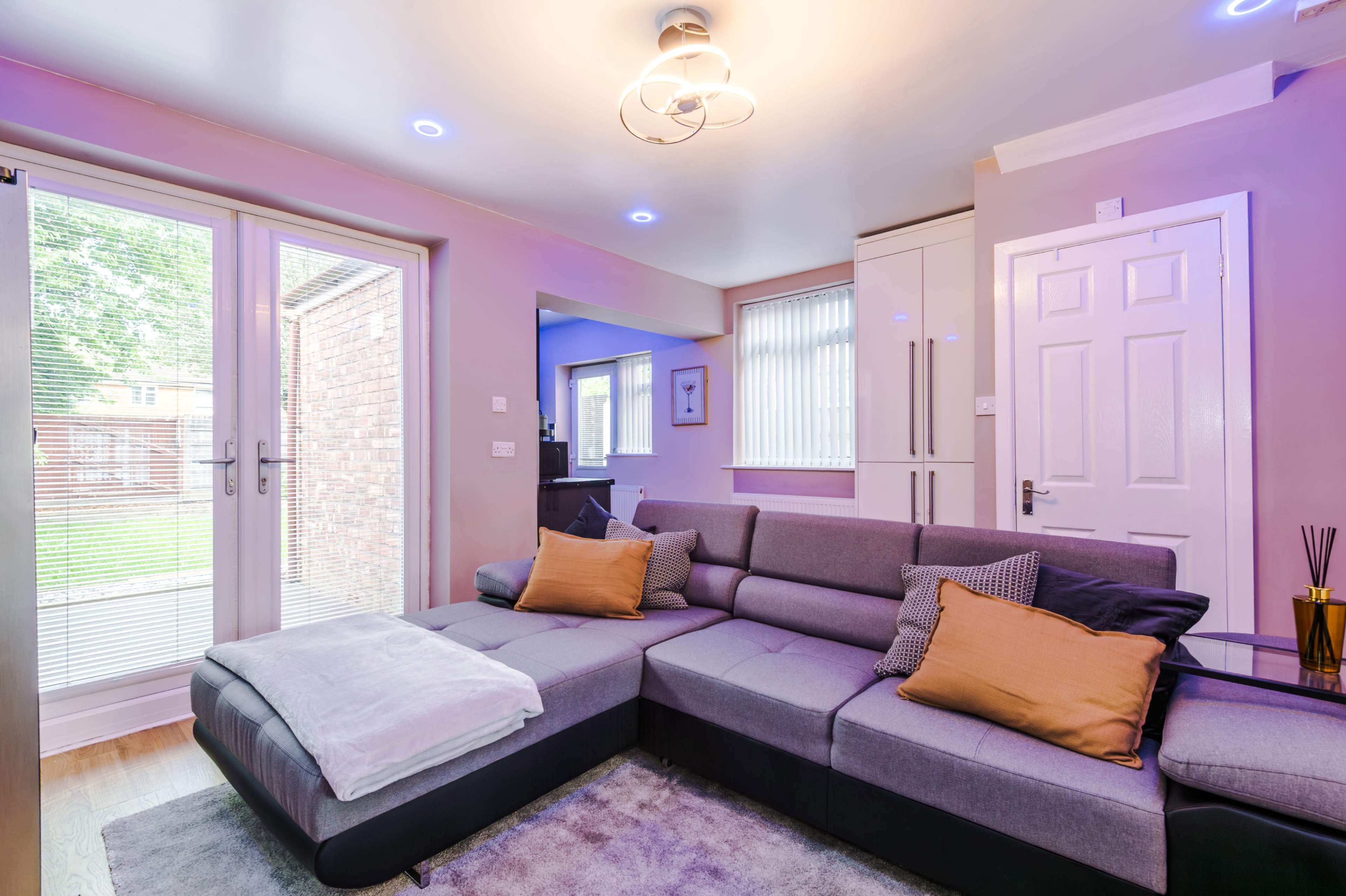 The image shows a modern living room with a gray sectional sofa, light fixtures, and large windows that open to a backyard.