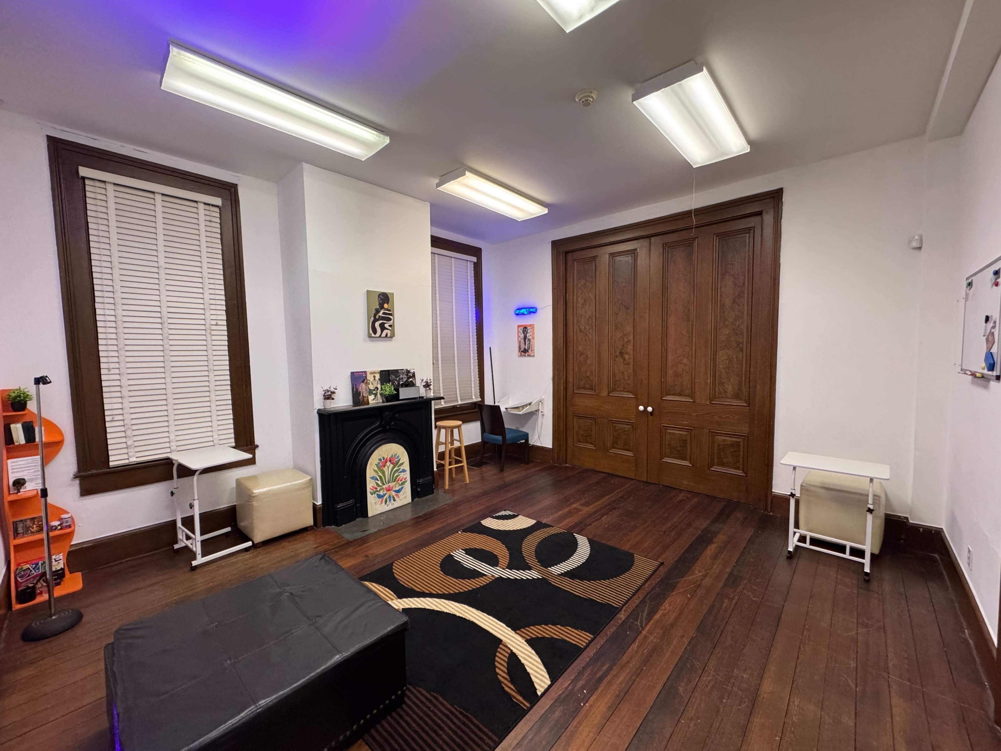 The image shows a room with wooden floors, a fireplace, and a mix of furniture including a black rug, a small table, and two white tables against the walls.