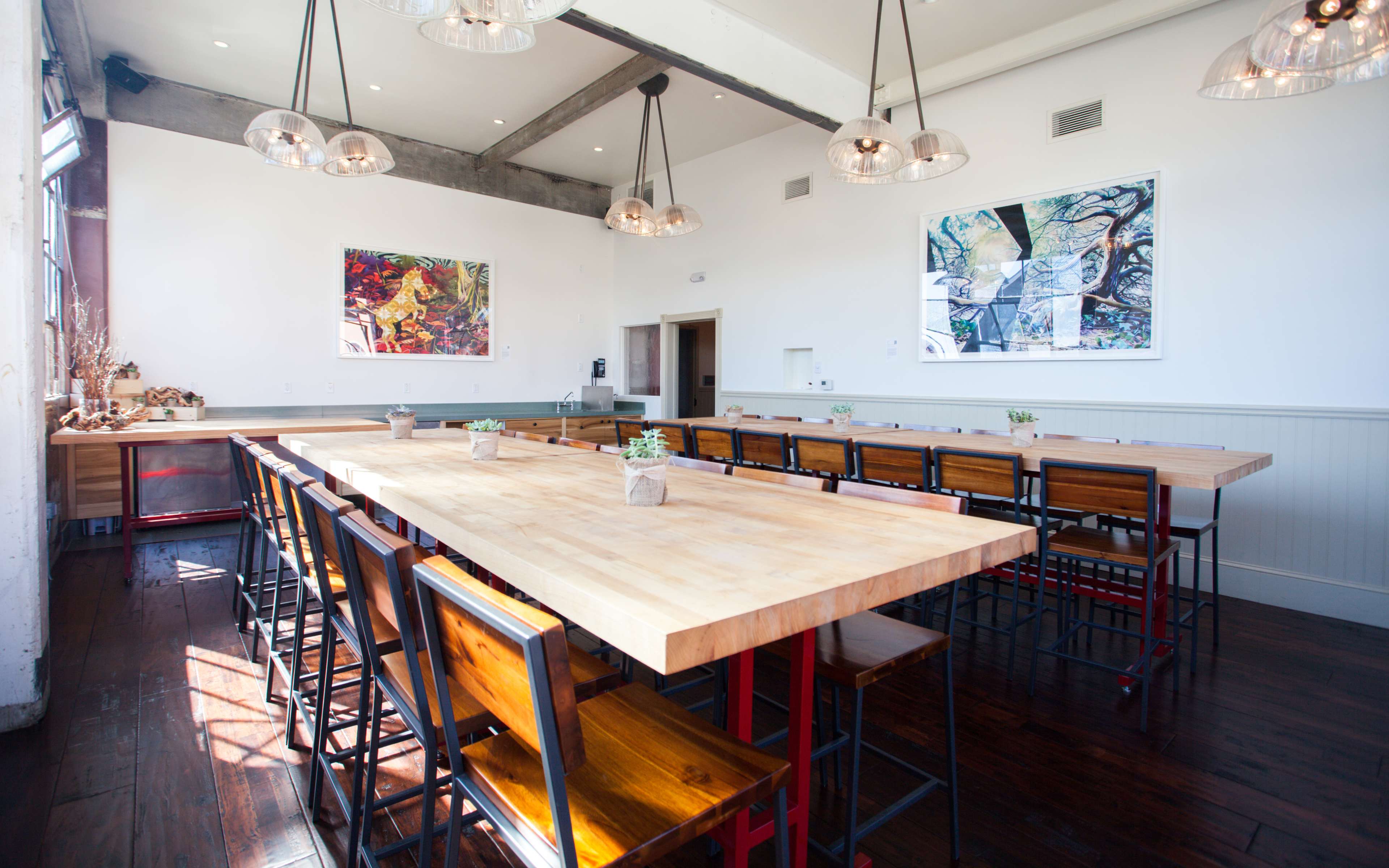The image shows a spacious dining area with a long wooden table surrounded by metal-framed chairs, adorned with small plants and featuring large windows that allow natural light to fill the room.