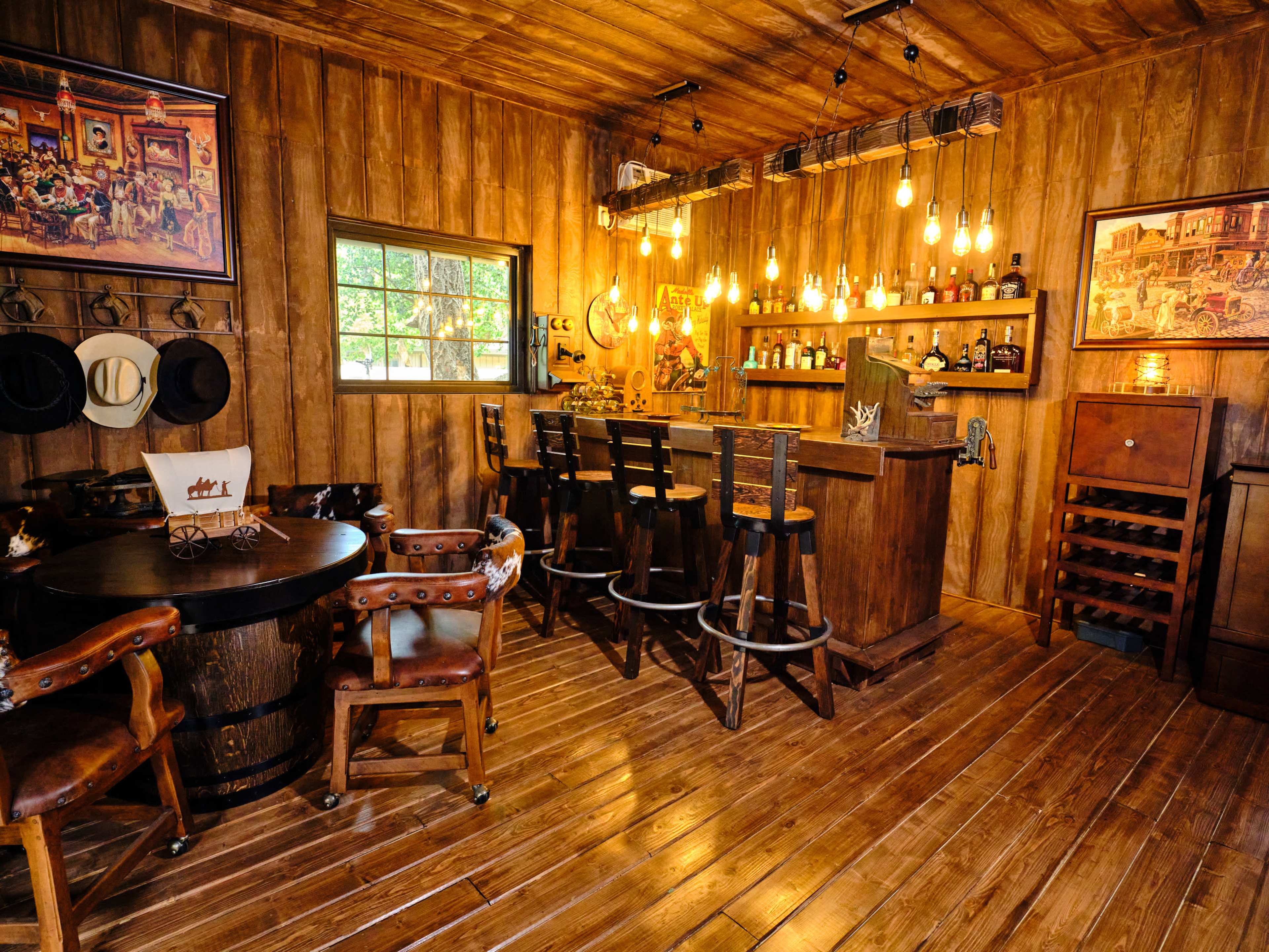 A rustic wooden bar area with a round table, chairs, and shelves lined with bottles, illuminated by pendant lights.