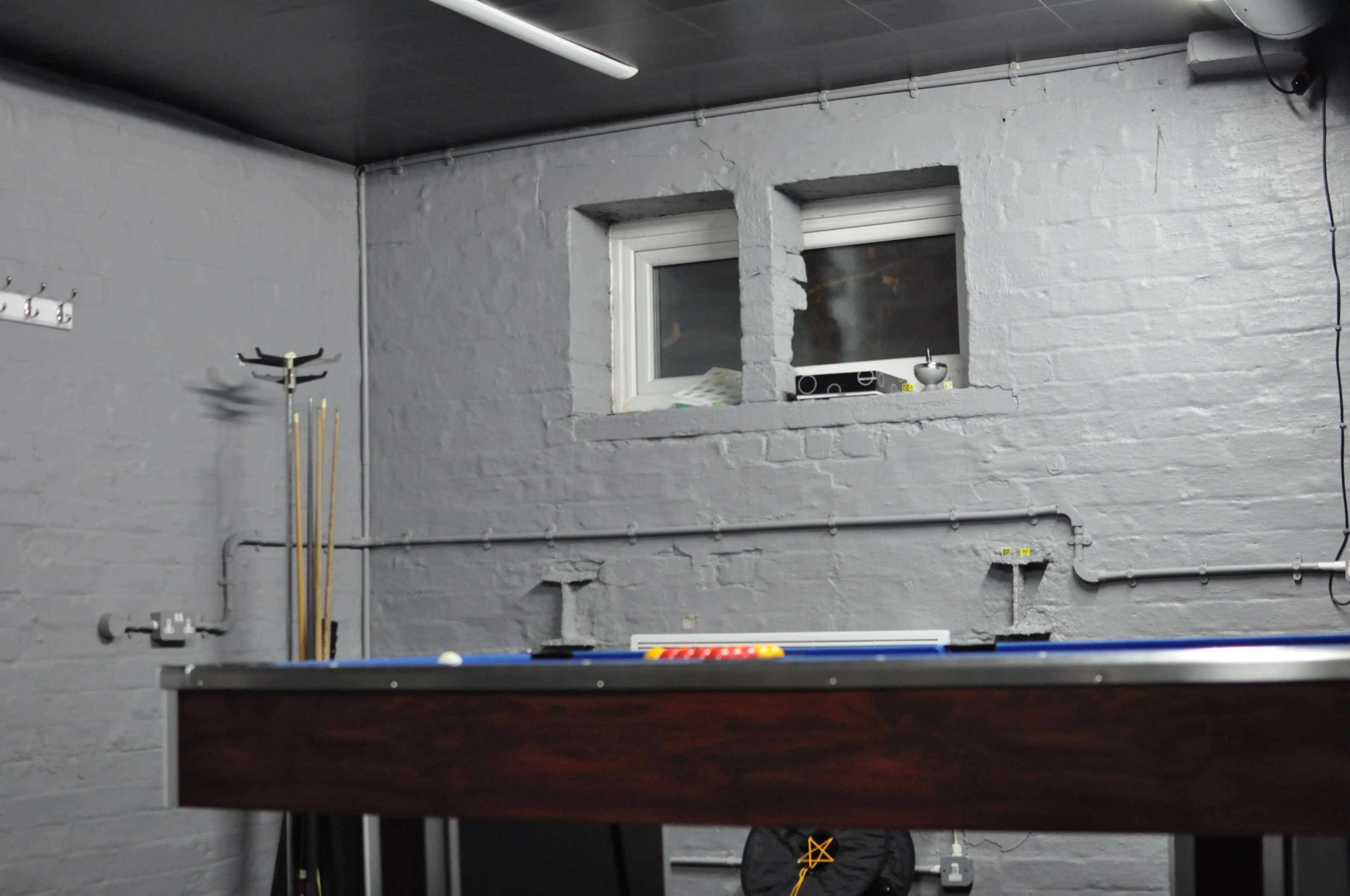 A billiards table is positioned in a dimly lit room with gray walls and a small window.
