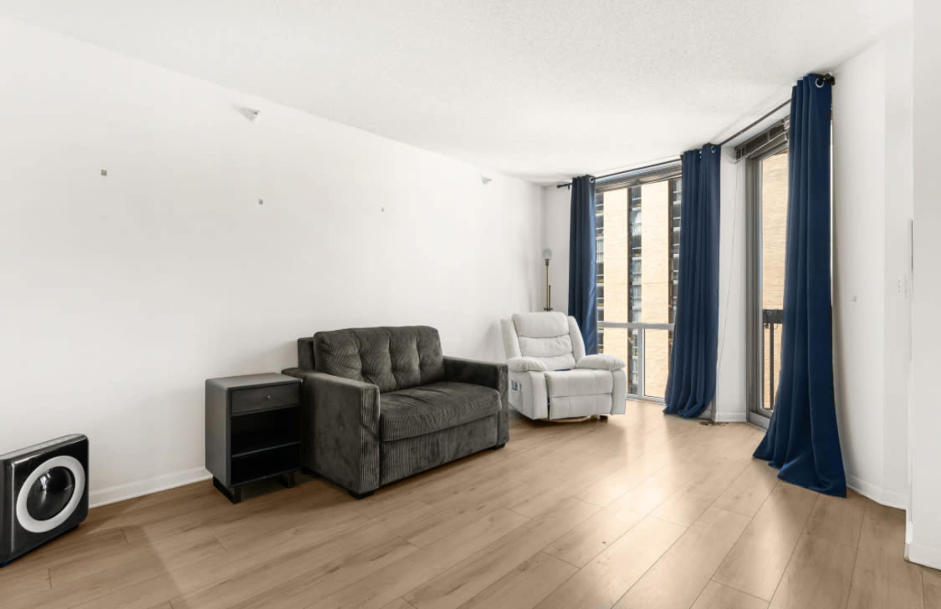 The image shows a spacious living room with a gray sofa, a white recliner, and large windows dressed with blue curtains.