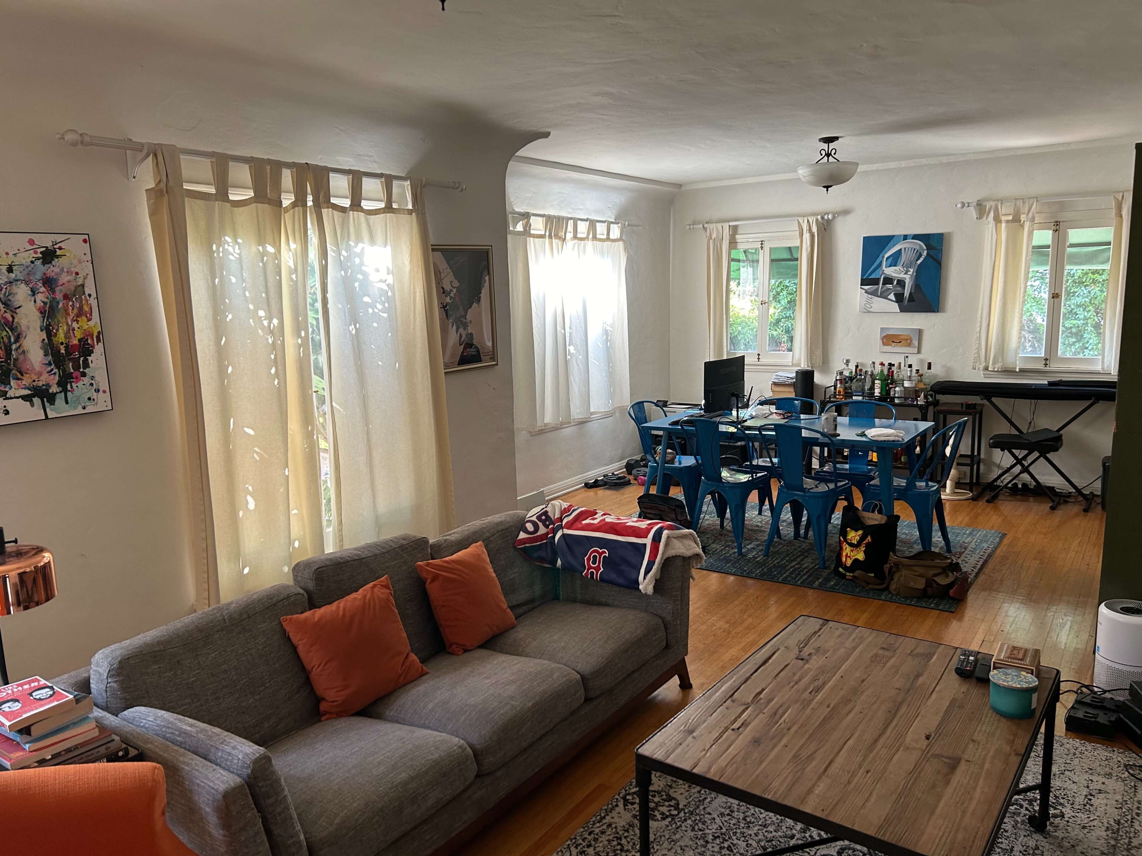 The image shows a living room with a gray sofa and orange cushions facing a dining area filled with blue chairs and a table, alongside a keyboard and various decorations.