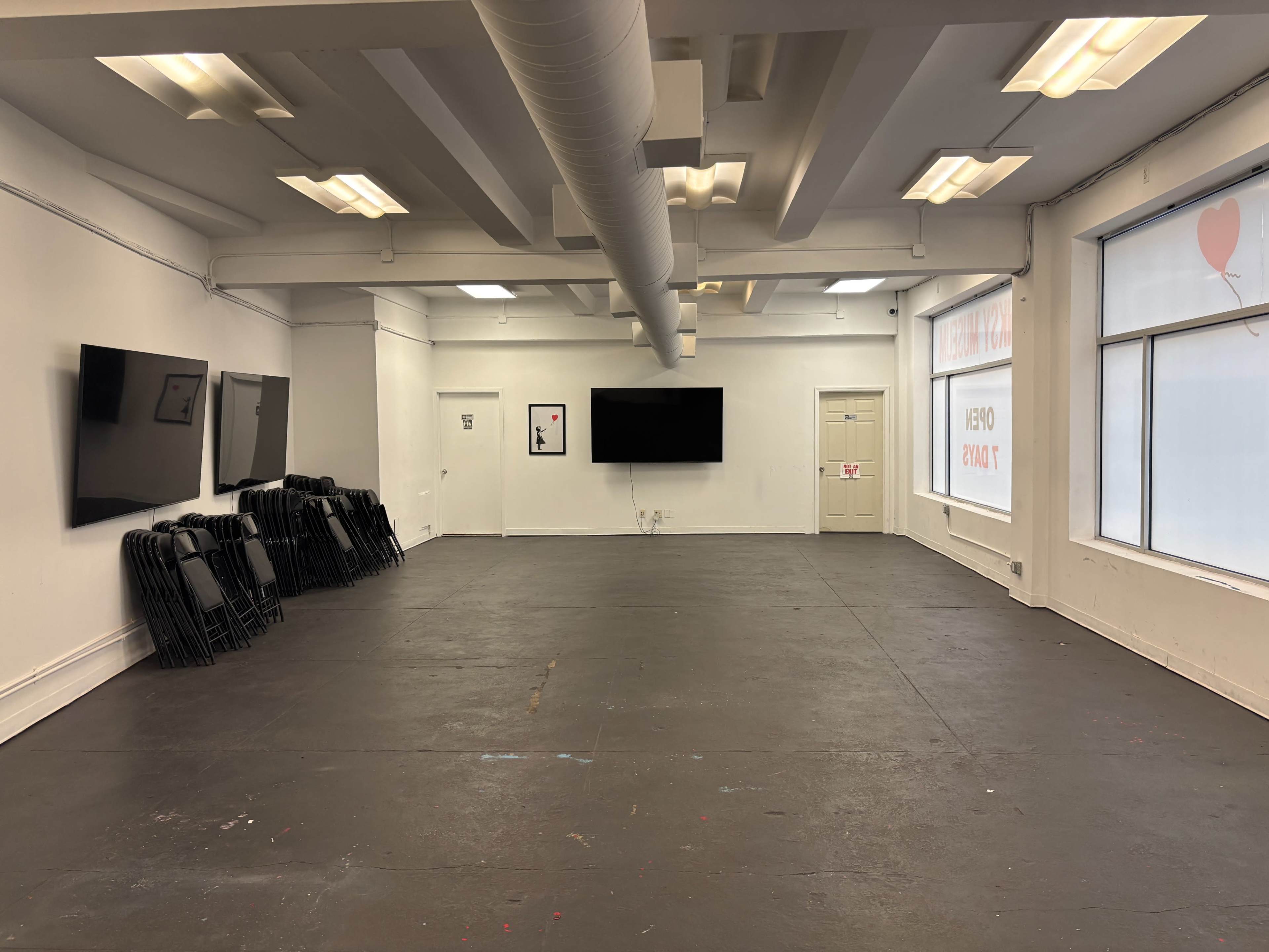 The image shows a spacious, empty room with several wall-mounted screens and a stack of black folding chairs in one corner.