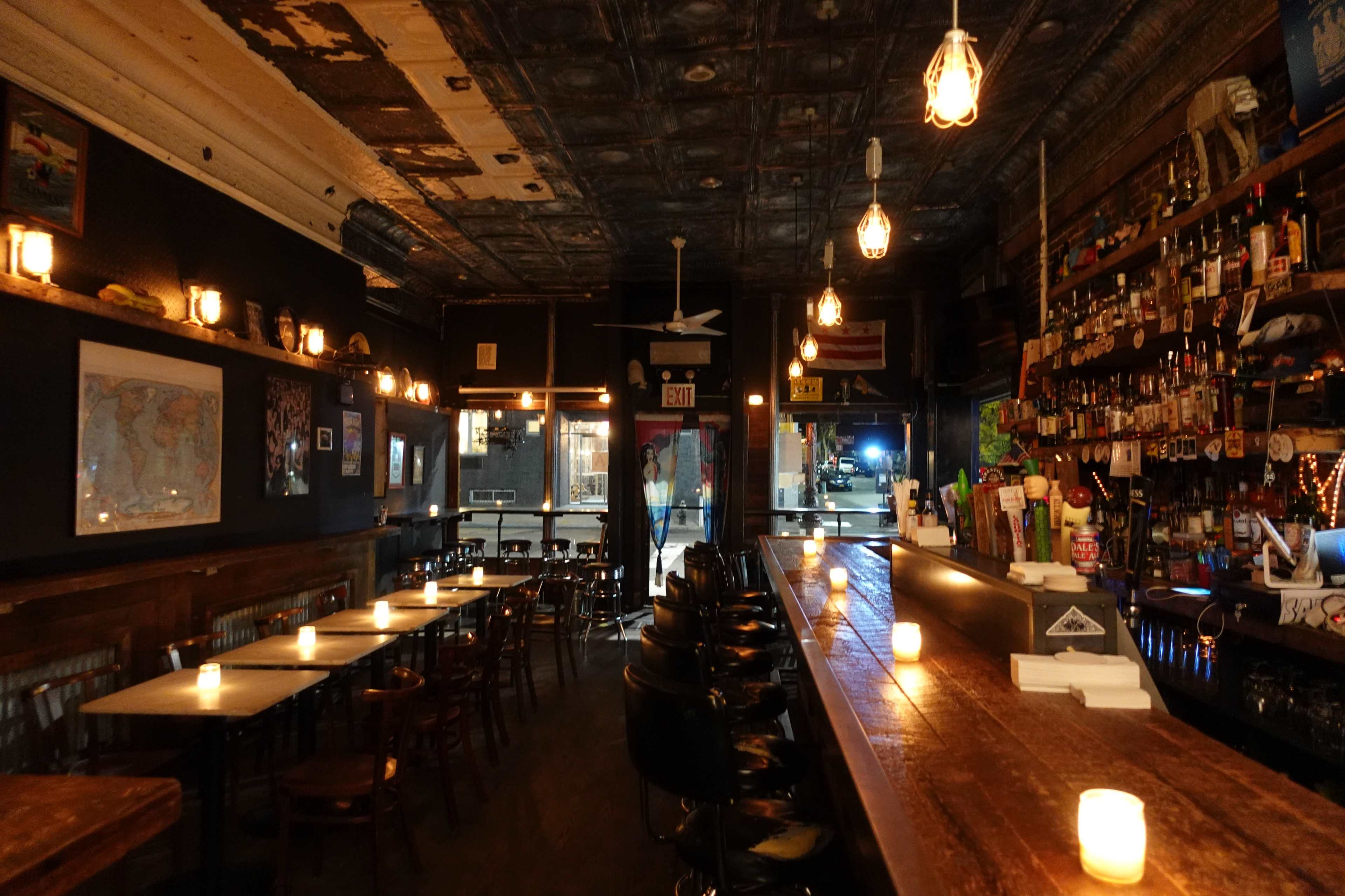 An empty bar with wooden tables and a long counter, illuminated by candles and pendant lights, set against a backdrop of a dimly lit interior featuring exposed ceiling tiles.