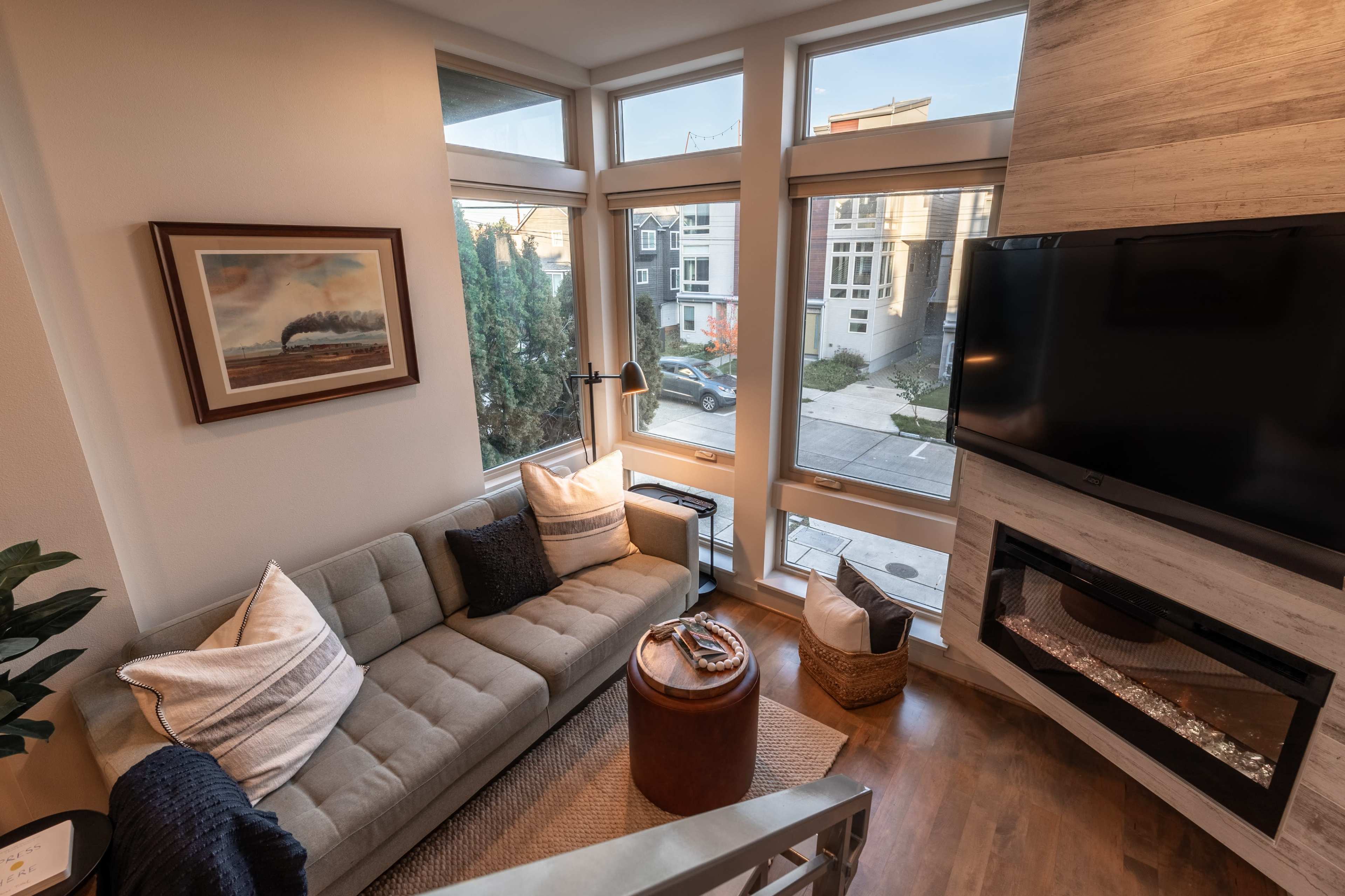 The image shows a modern living room with large windows, a sectional sofa, a television mounted on the wall, and a decorative fireplace.