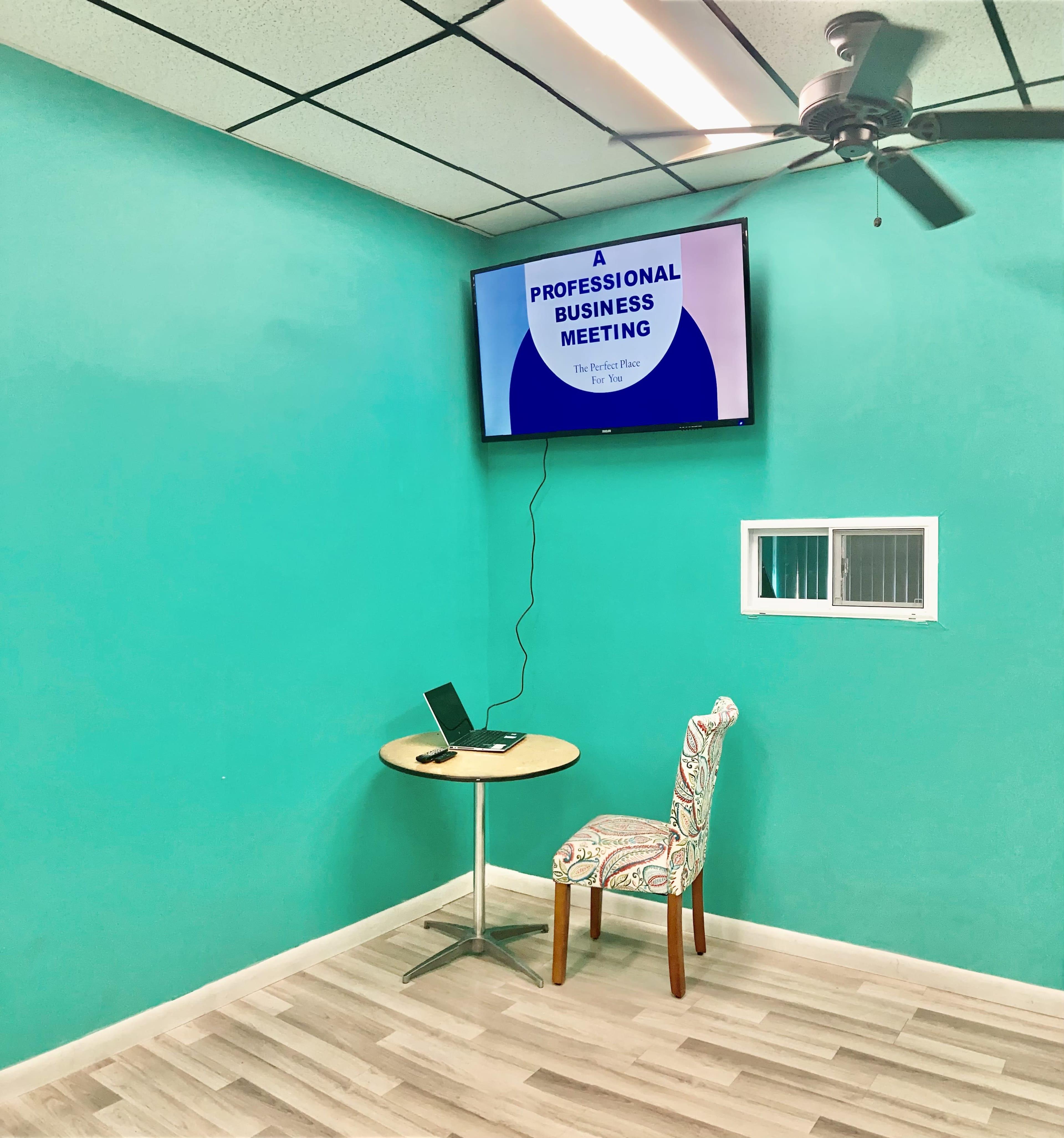 A small meeting room features a round table with a laptop, a patterned chair, and a wall-mounted screen displaying "A PROFESSIONAL BUSINESS MEETING."